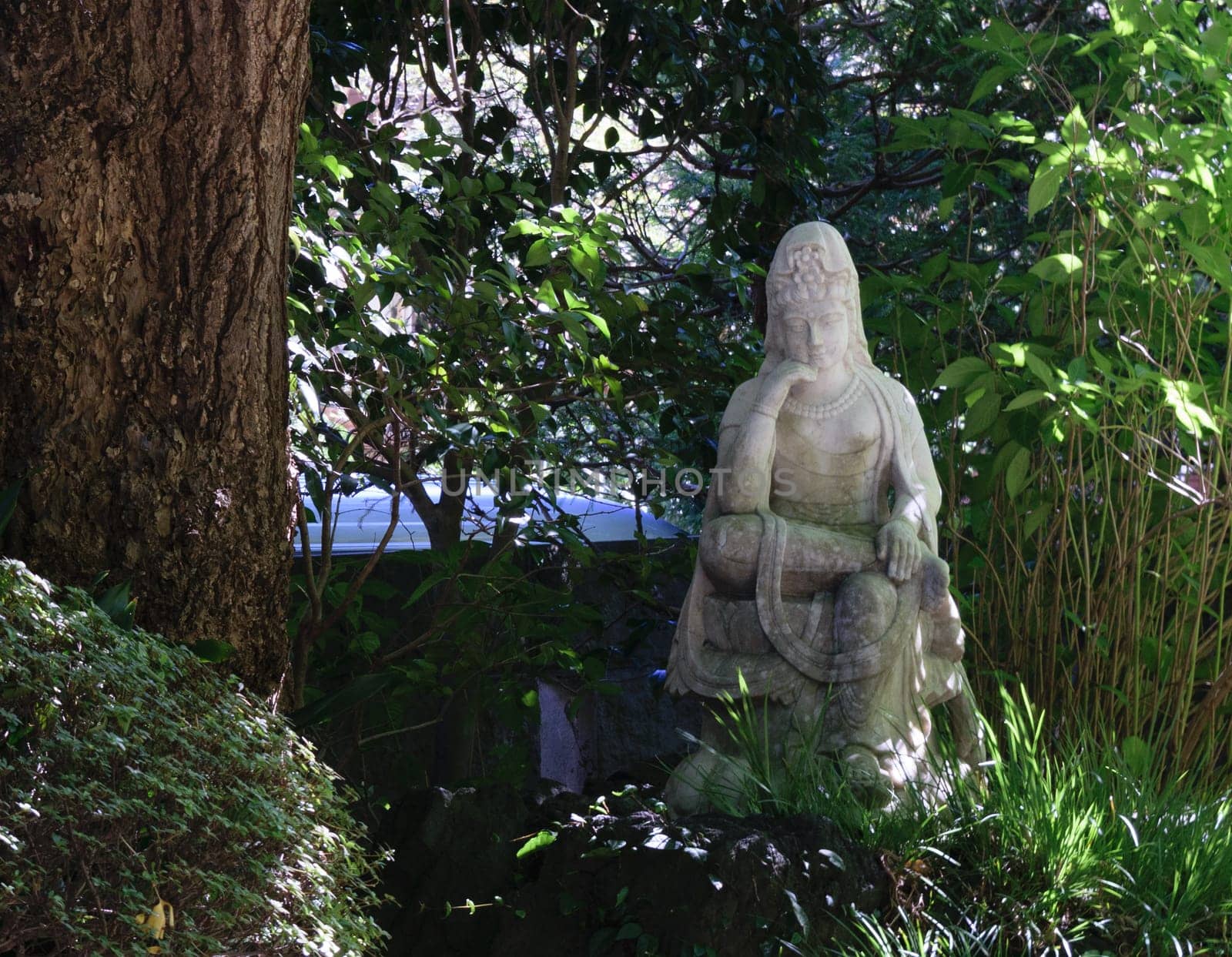 A stone statue of a seated figure surrounded by lush greenery and trees in a serene outdoor setting.