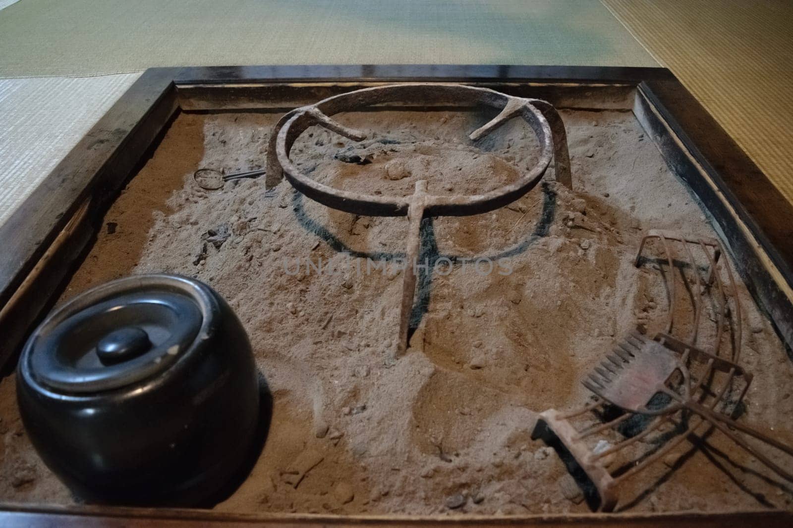 A traditional Japanese irori hearth with a cast iron pot and various tools on a sand-filled base. The hearth is surrounded by wooden frames and is used for cooking and heating.