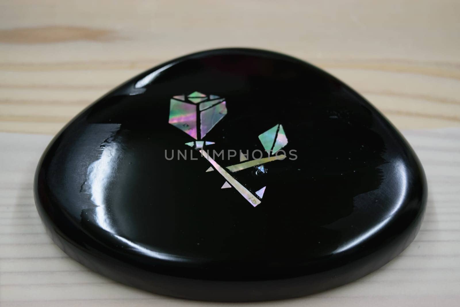 A smooth, black, polished stone with geometric inlay designs made of iridescent flower, placed on a light wooden surface.