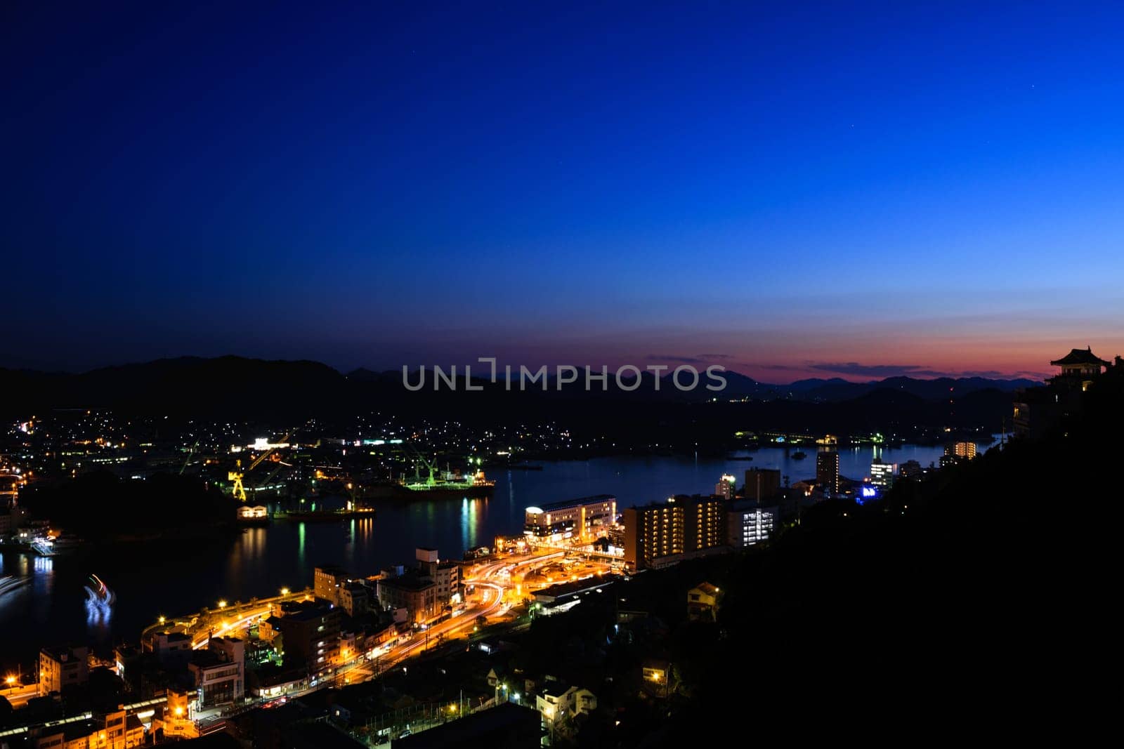 A cityscape at dusk with illuminated buildings and streets near a body of water. The sky transitions from deep blue to orange near the horizon.