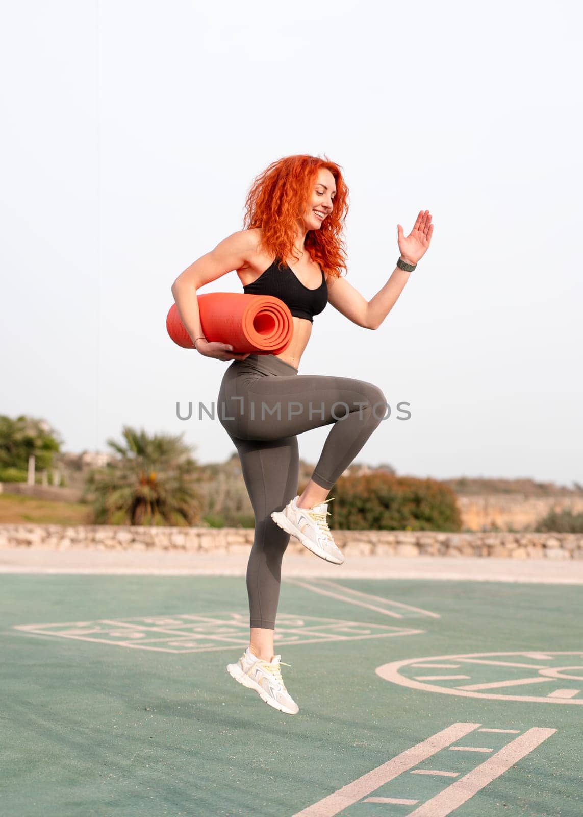 Active woman with redhead holding rolled mat doing warmup exercise. Happy beautiful young athlete doing jumping workout at park. Fitness and healthy lifestyles.