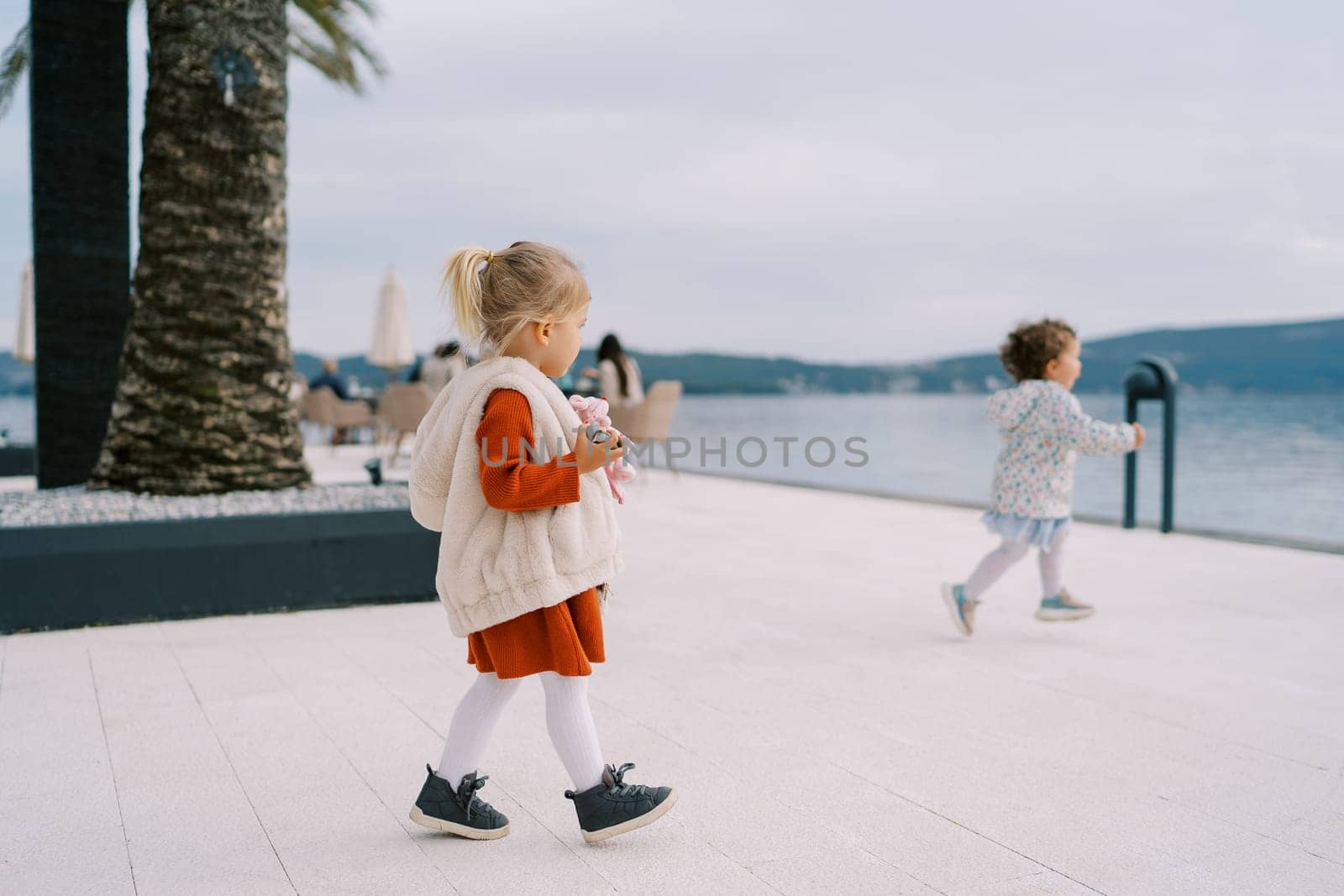 Little girl with a soft toy in her hand walks along the embankment towards the sea looking at the small girl running forward. High quality photo