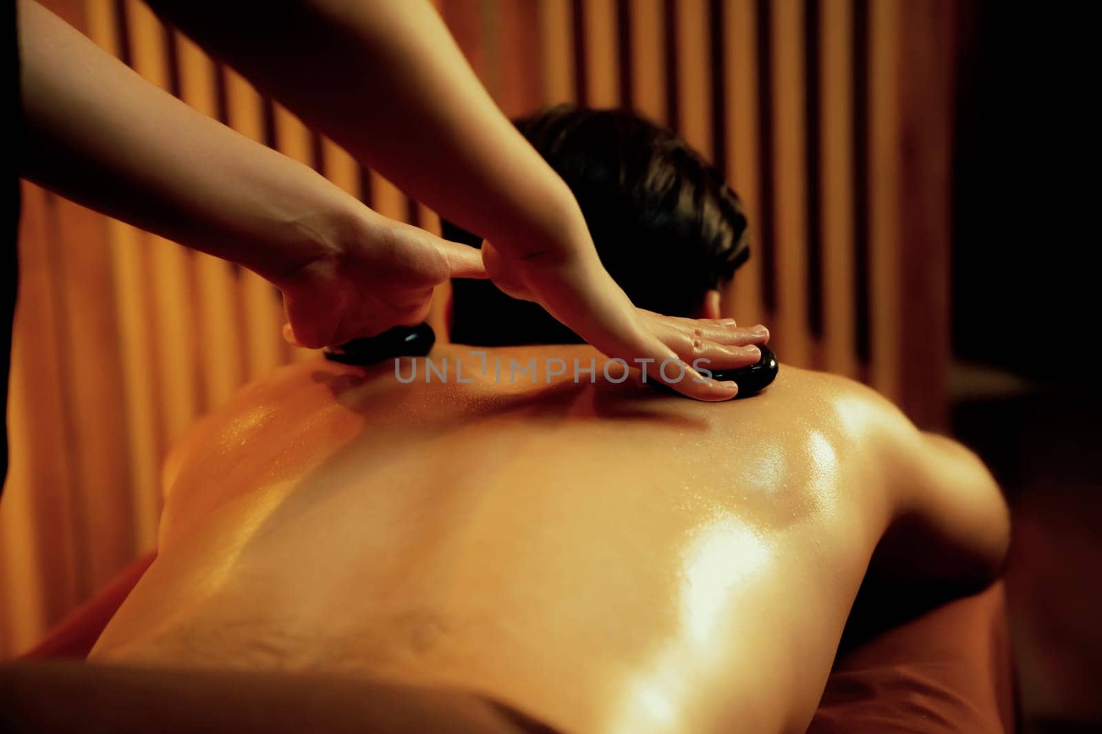Hot stone massage at spa salon in luxury resort with warm candle light, blissful man customer enjoying spa basalt stone massage glide over body with soothing warmth. Quiescent
