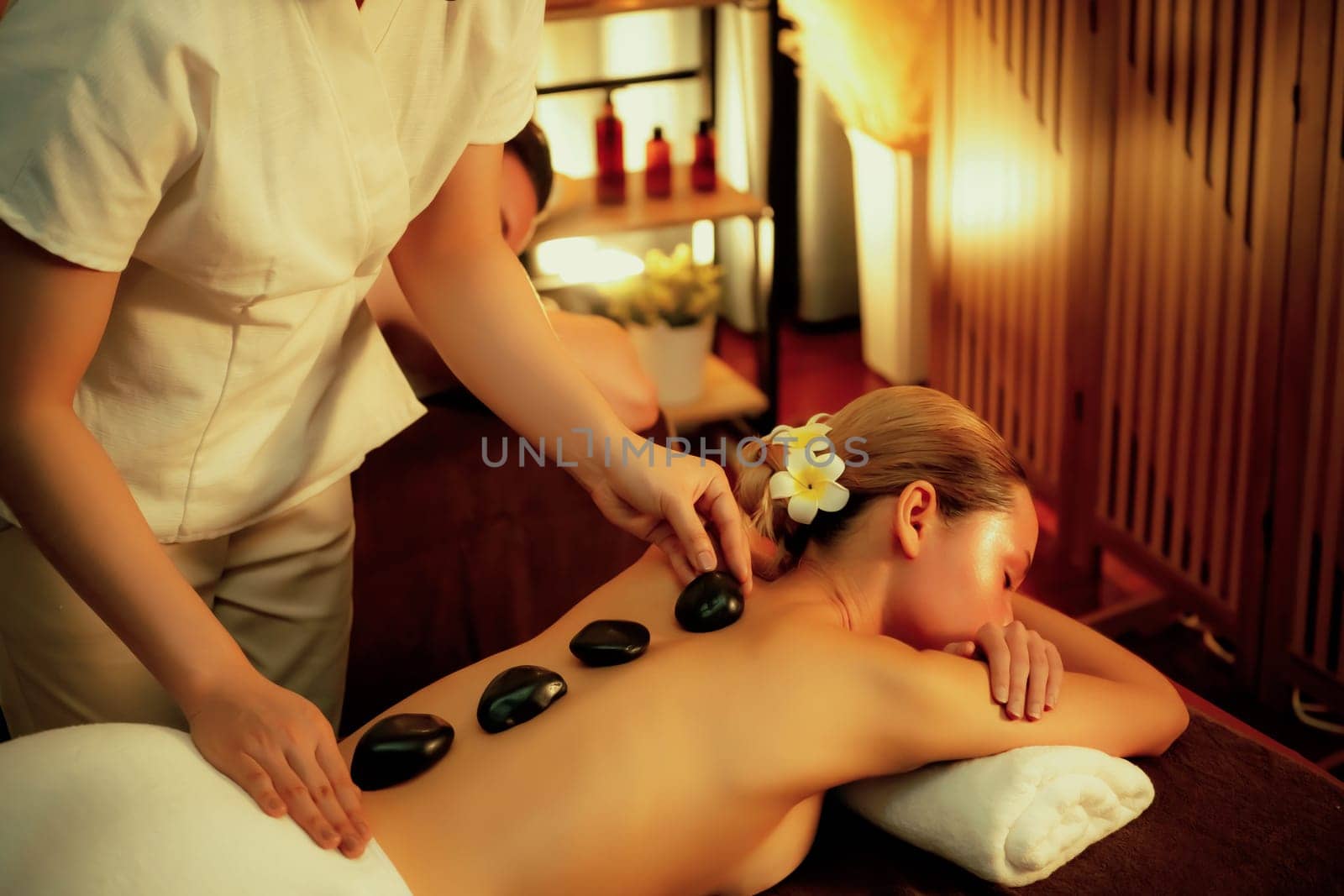 Hot stone massage at spa salon in luxury resort with warm candle light, blissful woman customer enjoying spa basalt stone massage glide over body with soothing warmth. Quiescent