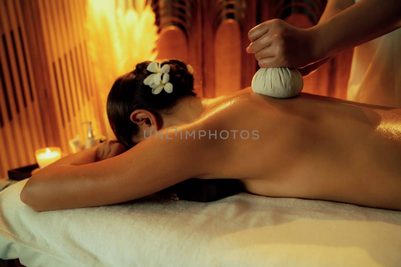 Hot herbal ball spa massage body treatment, masseur gently compresses herb bag on woman body. Tranquil and serenity of aromatherapy recreation in warm lighting of candles at spa salon. Quiescent