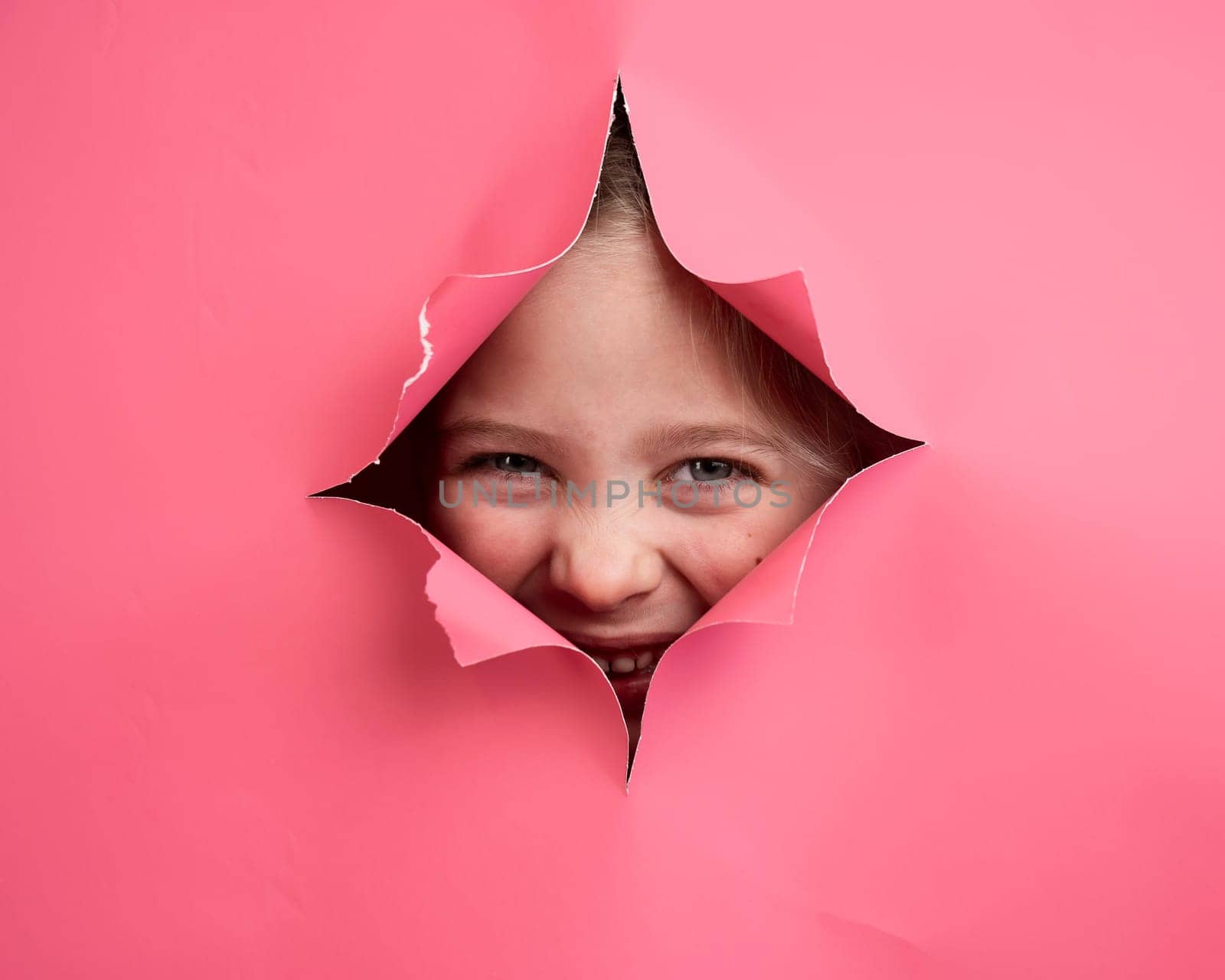 Cute Caucasian girl peeks out of a hole in a paper pink background