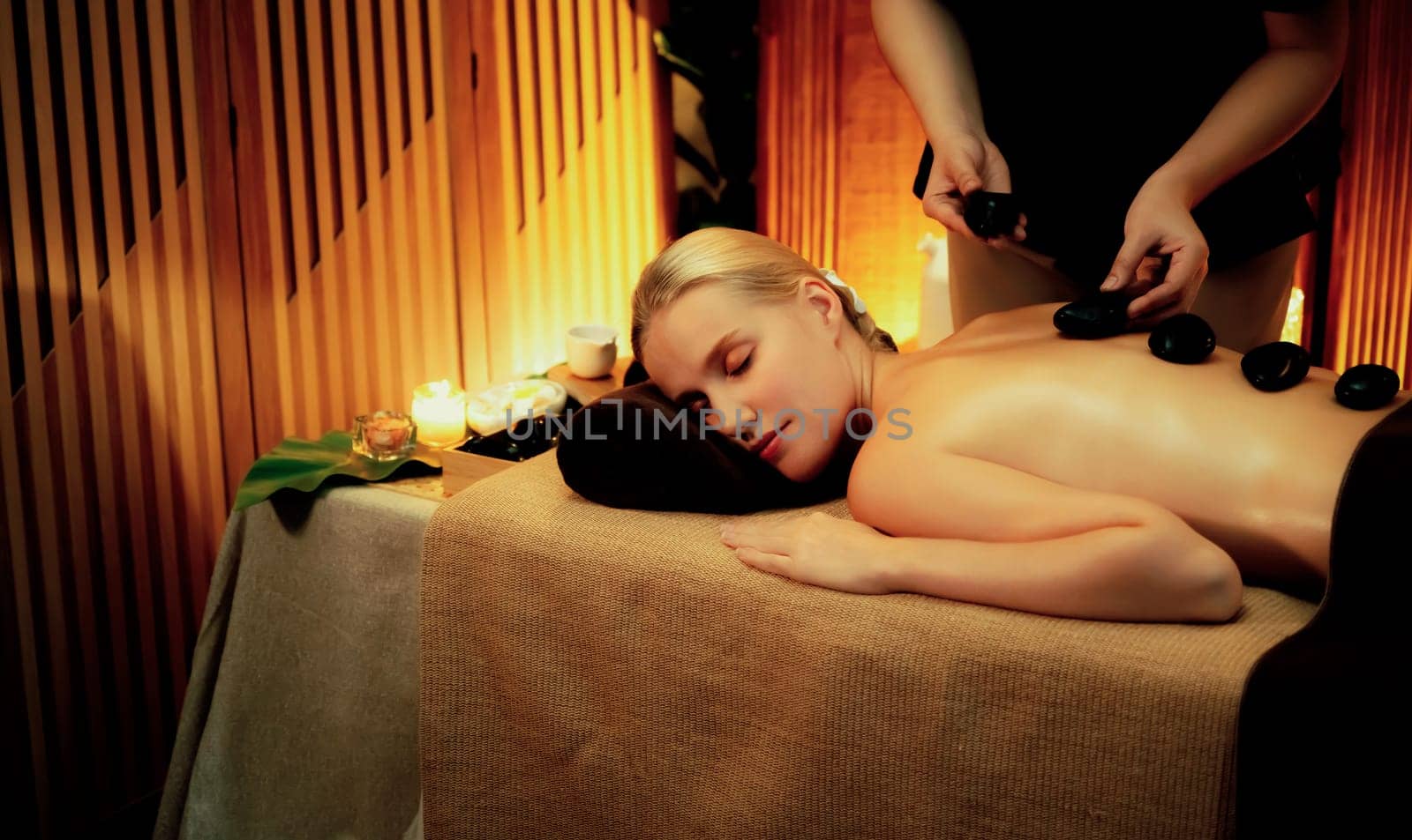 Hot stone massage at spa salon in luxury resort with warm candle light, blissful woman customer enjoying spa basalt stone massage glide over body with soothing warmth. Quiescent