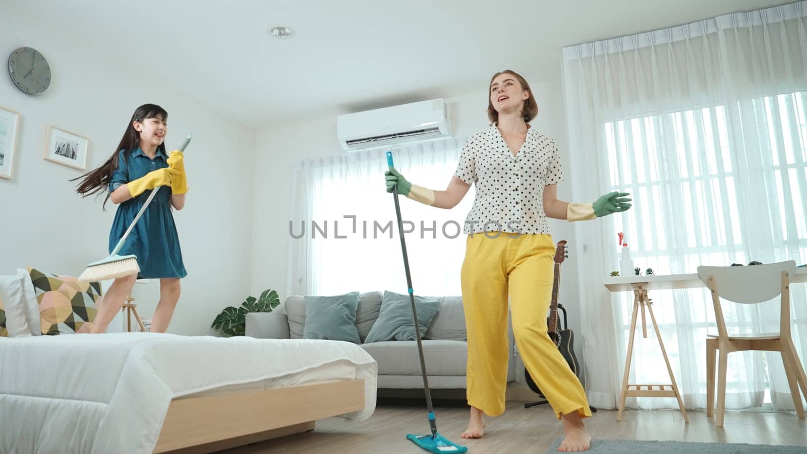 Caucasian mom and asian child doing housework together and dancing to music. Attractive mother and daughter cleaning modern bedroom while sing along and jump to relaxing music. Lifestyle. Pedagogy.