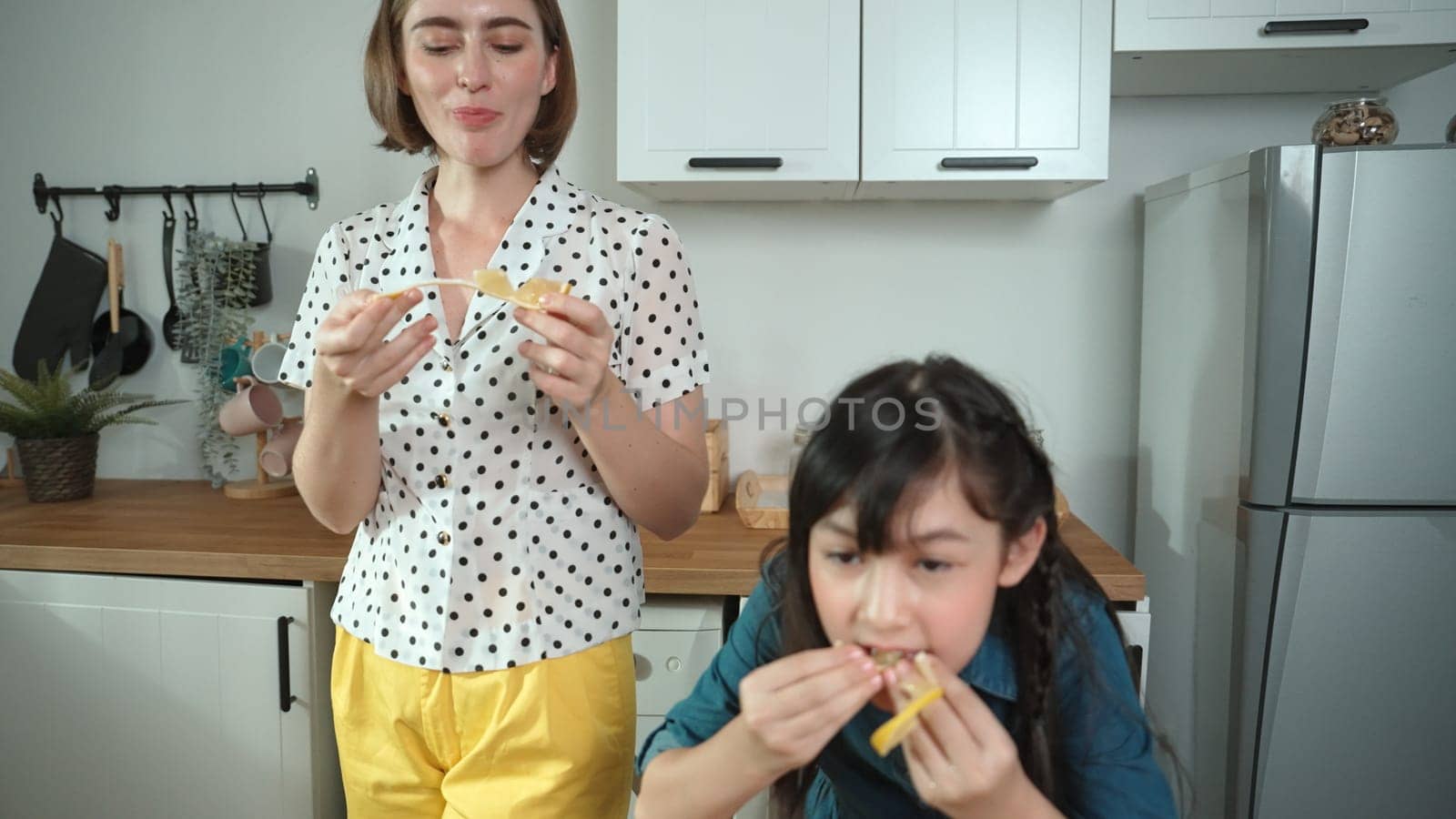 Energetic caucasian mother and asian girl cooking together and eating orange or preparing salad for dinner. Happy mom and daughter making healthy food with fresh food. Healthy food concept. Pedagogy.