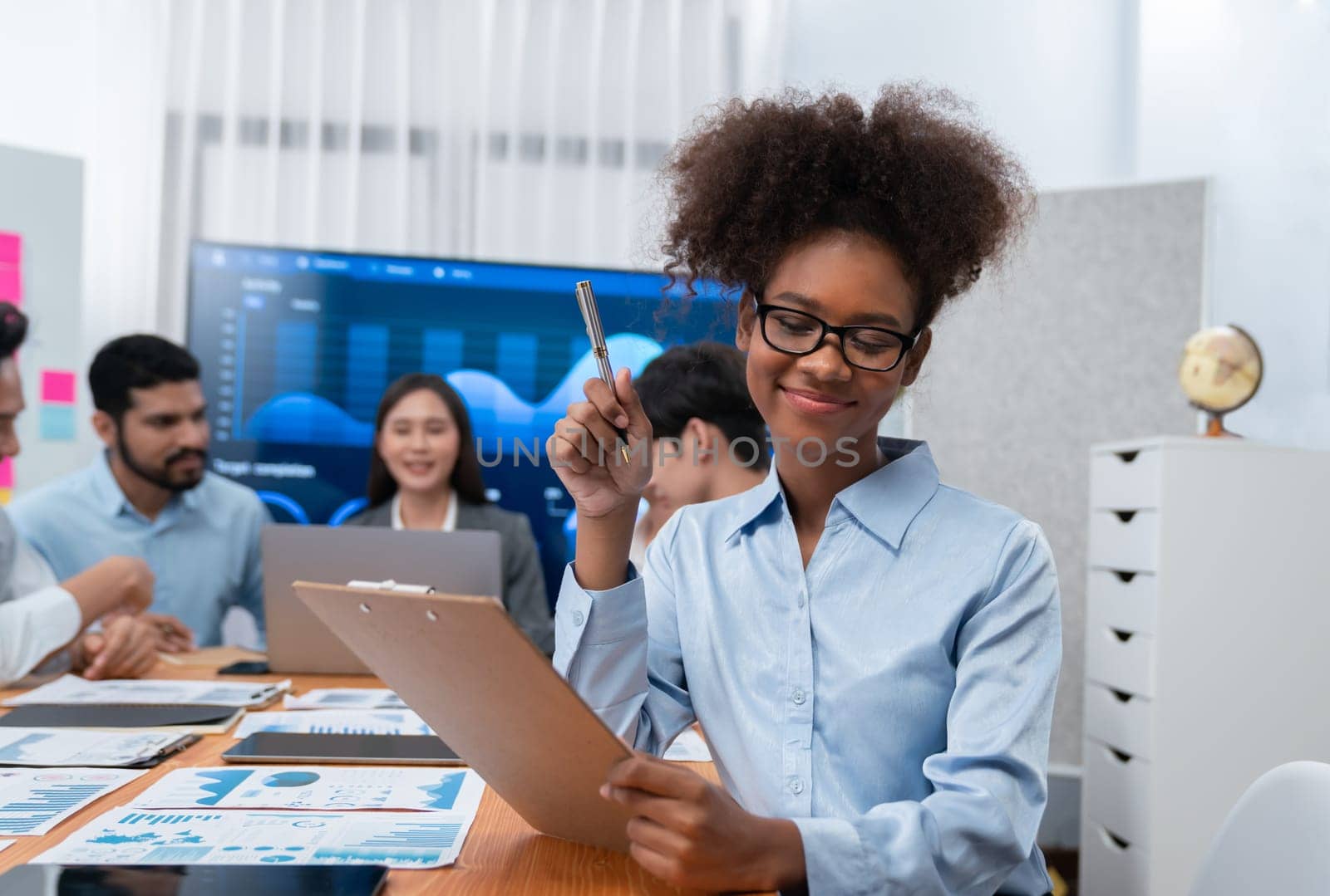 Happy young african businesswoman wearing glasses portrait with group of office worker on meeting with screen display business dashboard in background. Confident office lady at team meeting. Concord