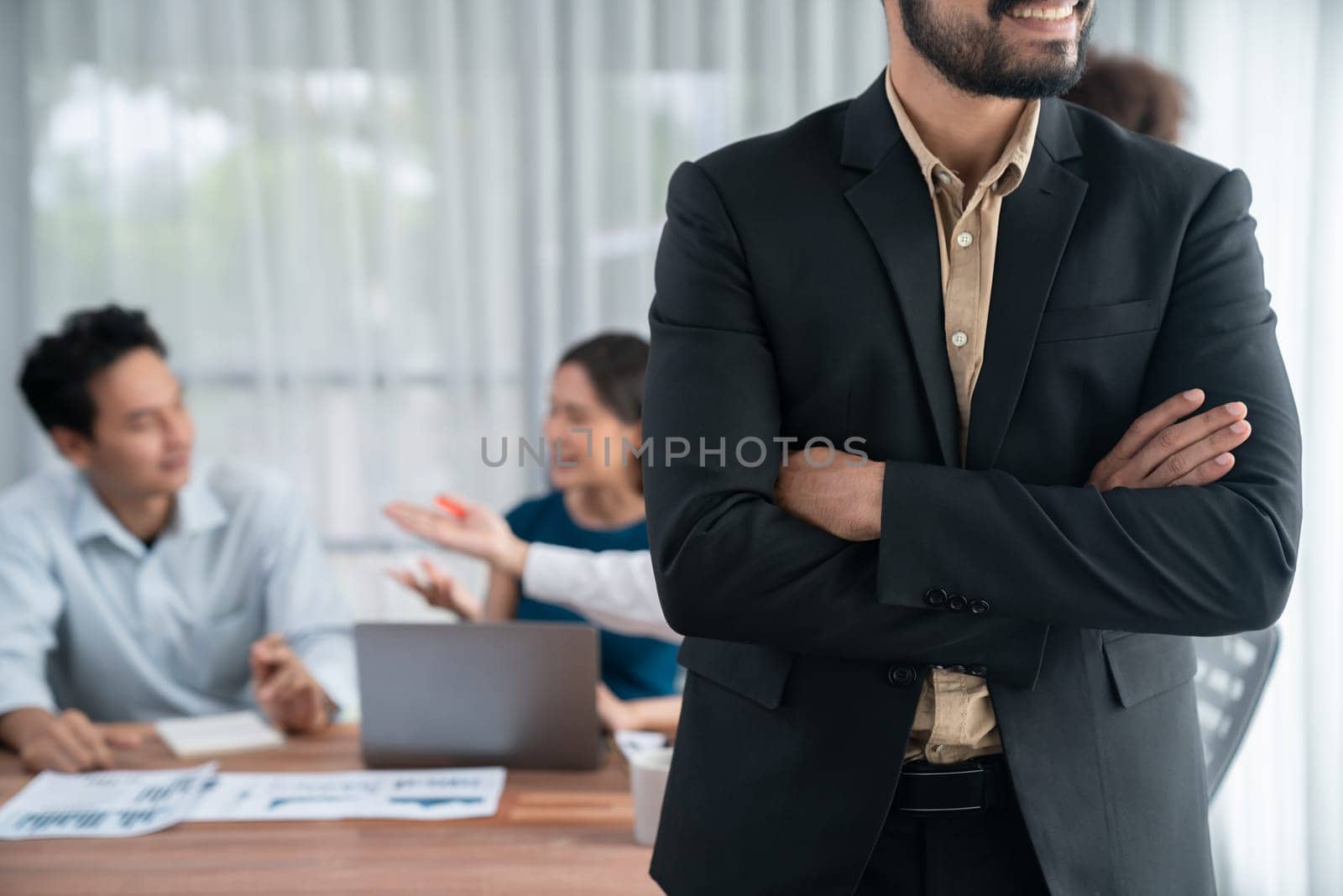 Businessman poses confidently with diverse coworkers in busy meeting room background. Multicultural team works together for business success. Modern businessman portrait. Concord