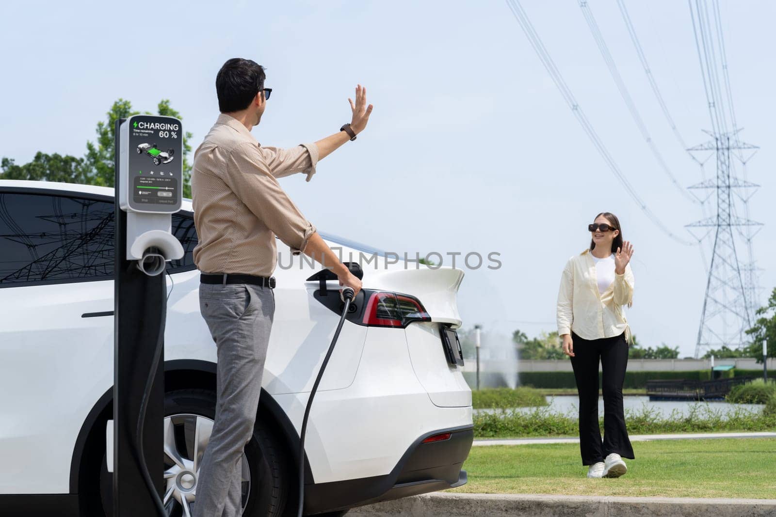 Young couple recharge EV car battery at charging station connected to power grid tower electrical industrial facility as electrical industry for eco friendly vehicle utilization. Expedient