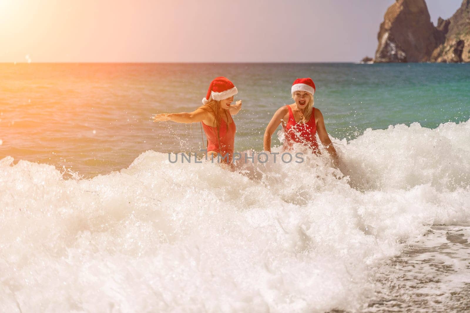 Women Santa hats ocean play. Seaside, beach daytime, enjoying beach fun. Two women in red swimsuits and Santa hats are enjoying themselves in the ocean waves