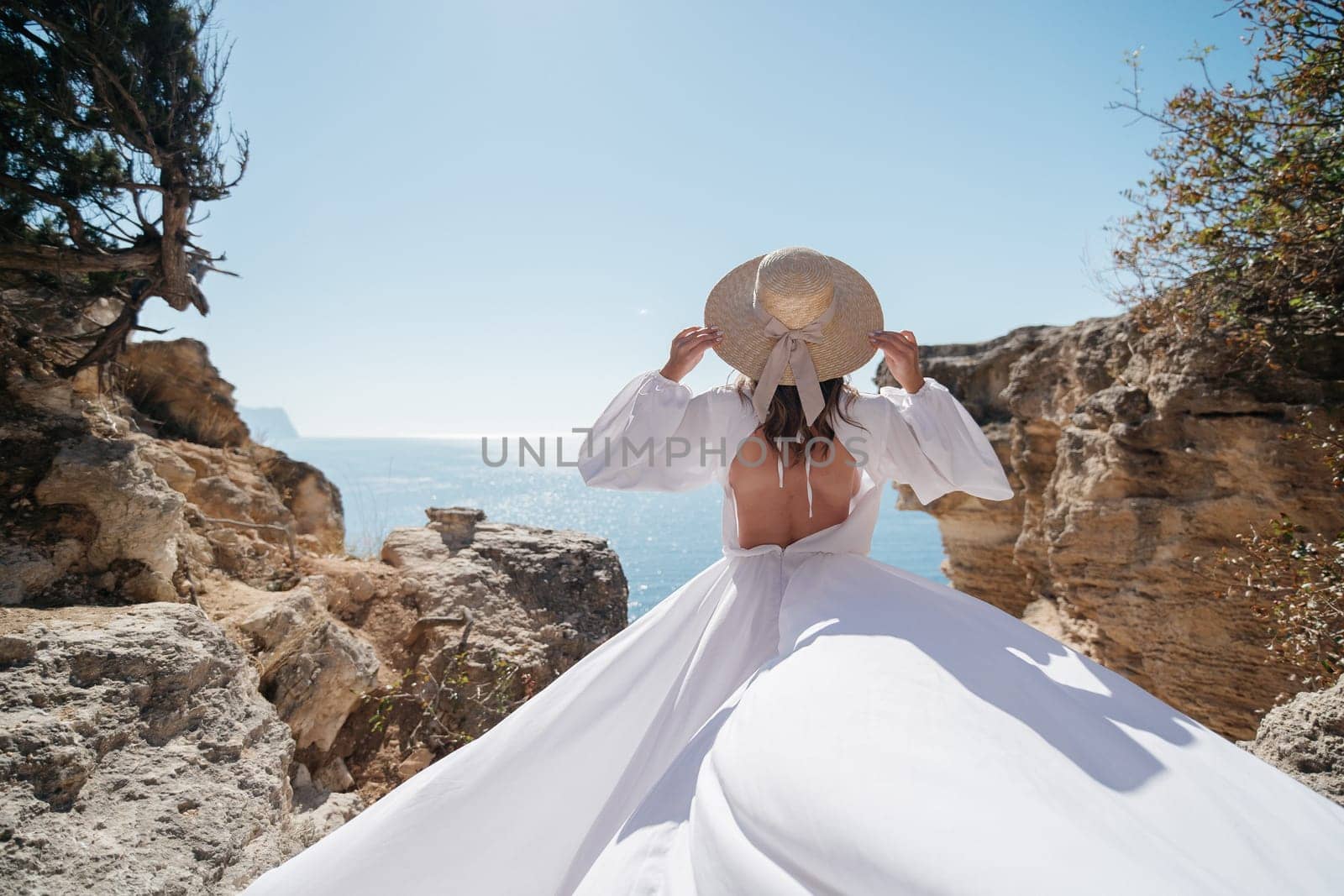 A woman in a white dress is standing on a rocky cliff overlooking the ocean. She is wearing a straw hat and she is enjoying the view