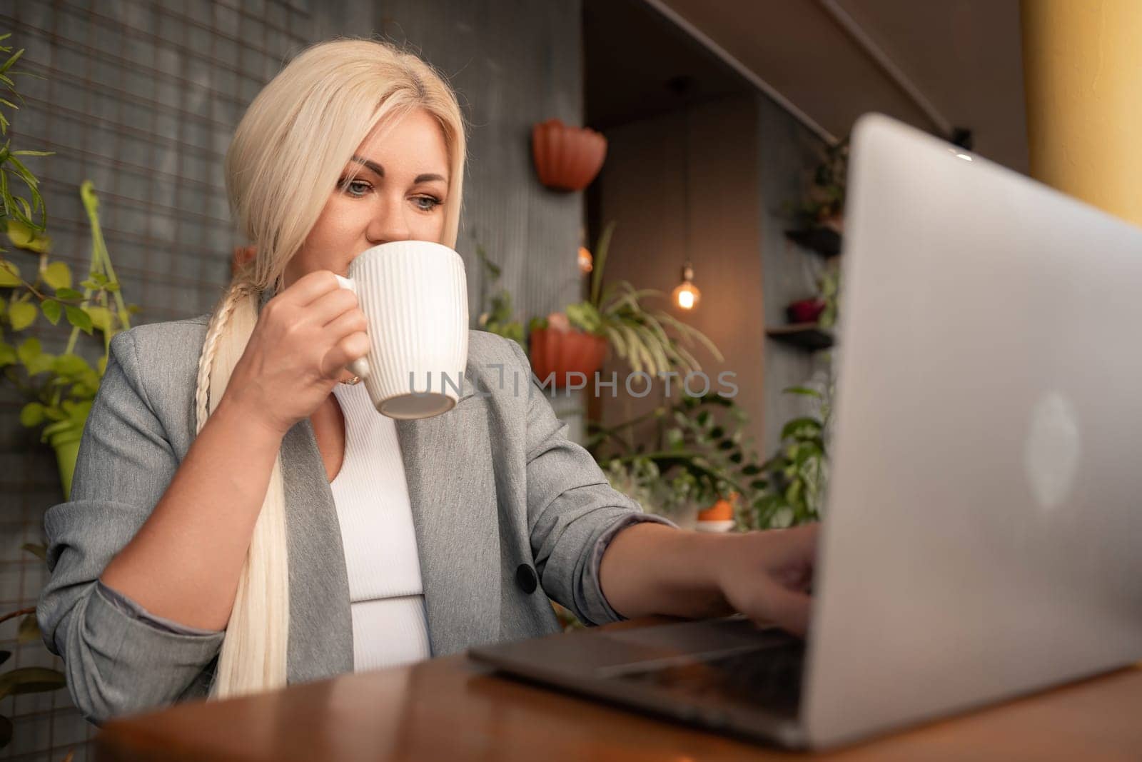 A woman is sitting at a table with a laptop and a white coffee cup. She is drinking coffee while working on her laptop