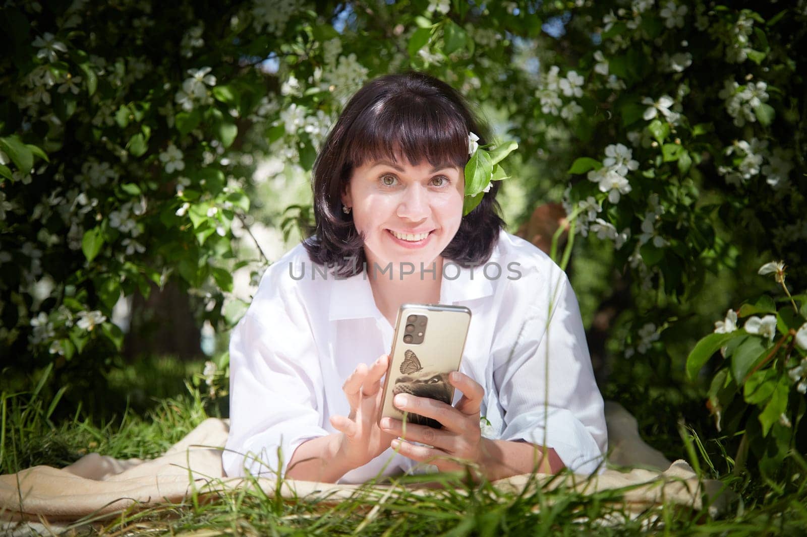 Brunette girl Using Smartphone in Blossoming Orchard in Springtime. Middle aged Woman enjoying phone among spring blossoms of apple or sakura trees