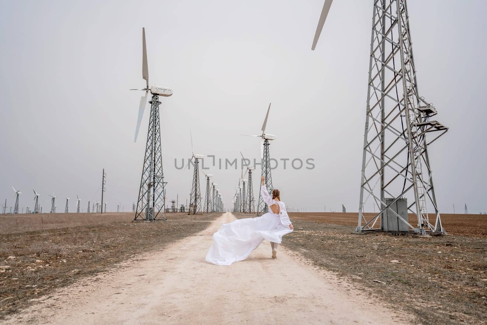 A woman in a white dress is walking down a dirt road in front of a row of wind turbines