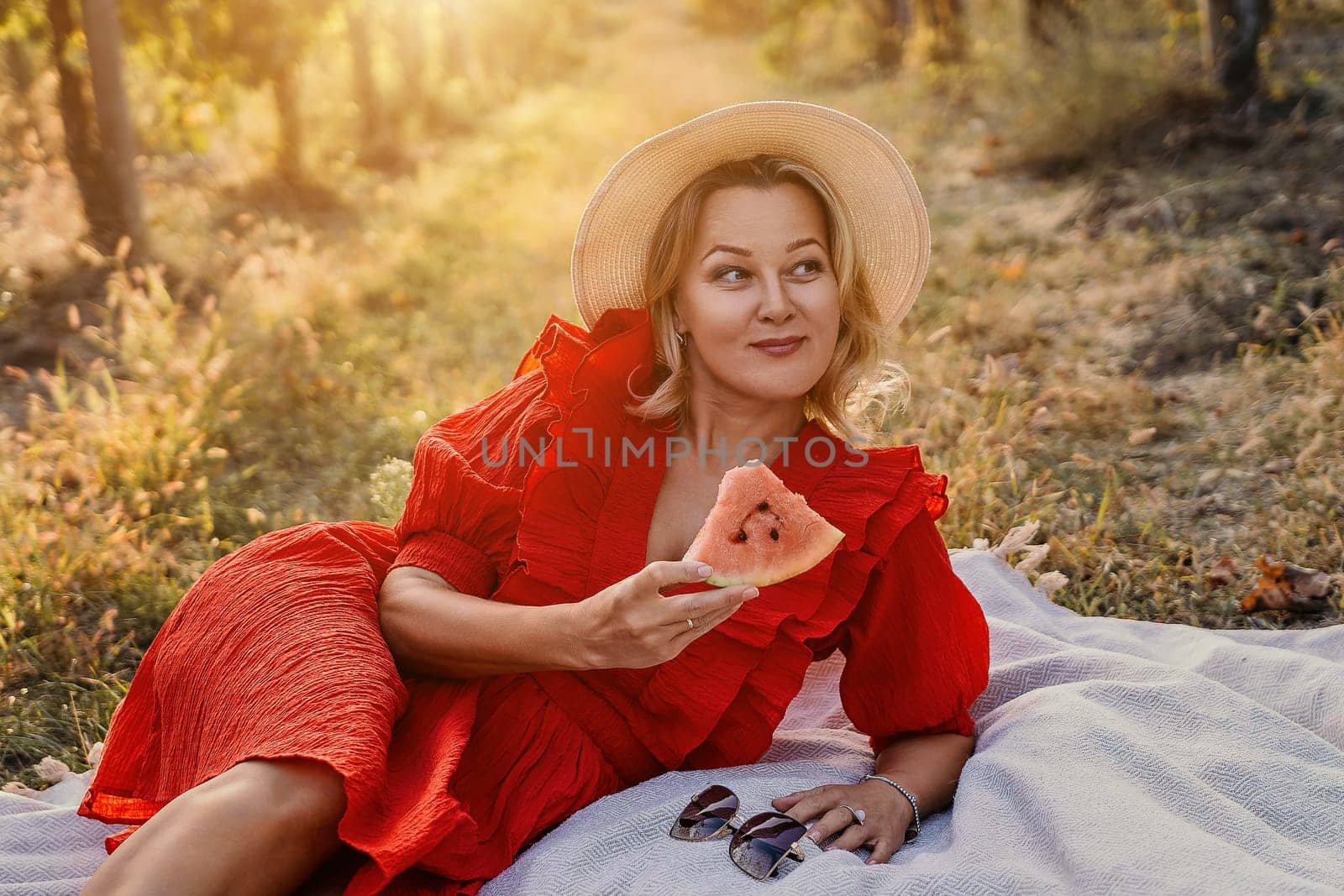 A woman in a red dress is laying on a blanket and holding a slice of watermelon. The scene is peaceful and relaxing, with the woman enjoying a simple moment outdoors