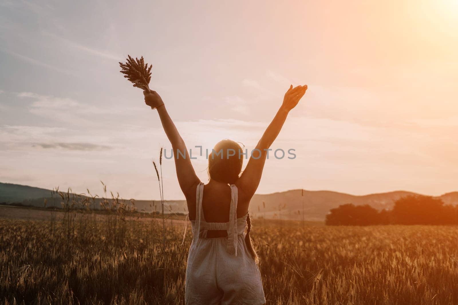 A woman is holding a bunch of wheat in her arms. The wheat is dry and brown, and the woman is wearing a white dress. The scene is set in a field, and the woman is posing for a photo