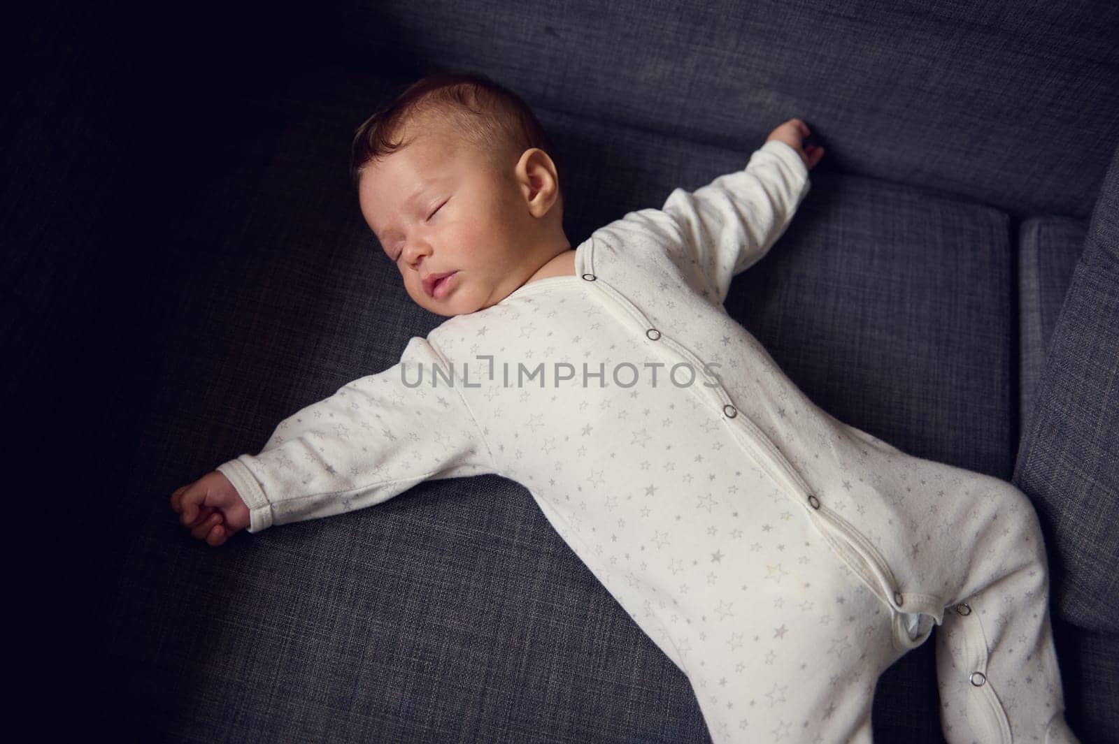 View from above of a cute newborn baby boy in white bodysuit, sweetly sleeping on comfortable gray sofa. European lovely child baby boy 4-6 months old, sleeping with his outstretched hands