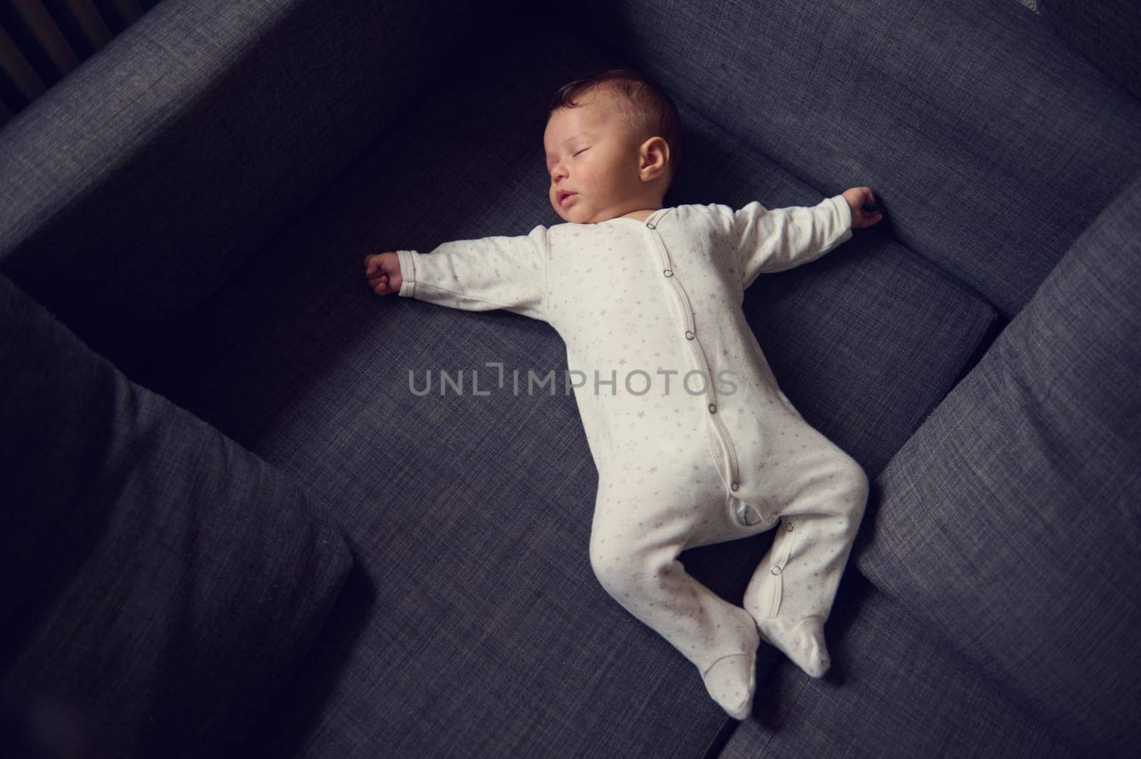 Authentic portrait of European cute baby boy in white bodysuit, sweetly sleeping on comfortable gray sofa. The concept of happy childhood. Children. Infancy and babyhood. World Children's Day