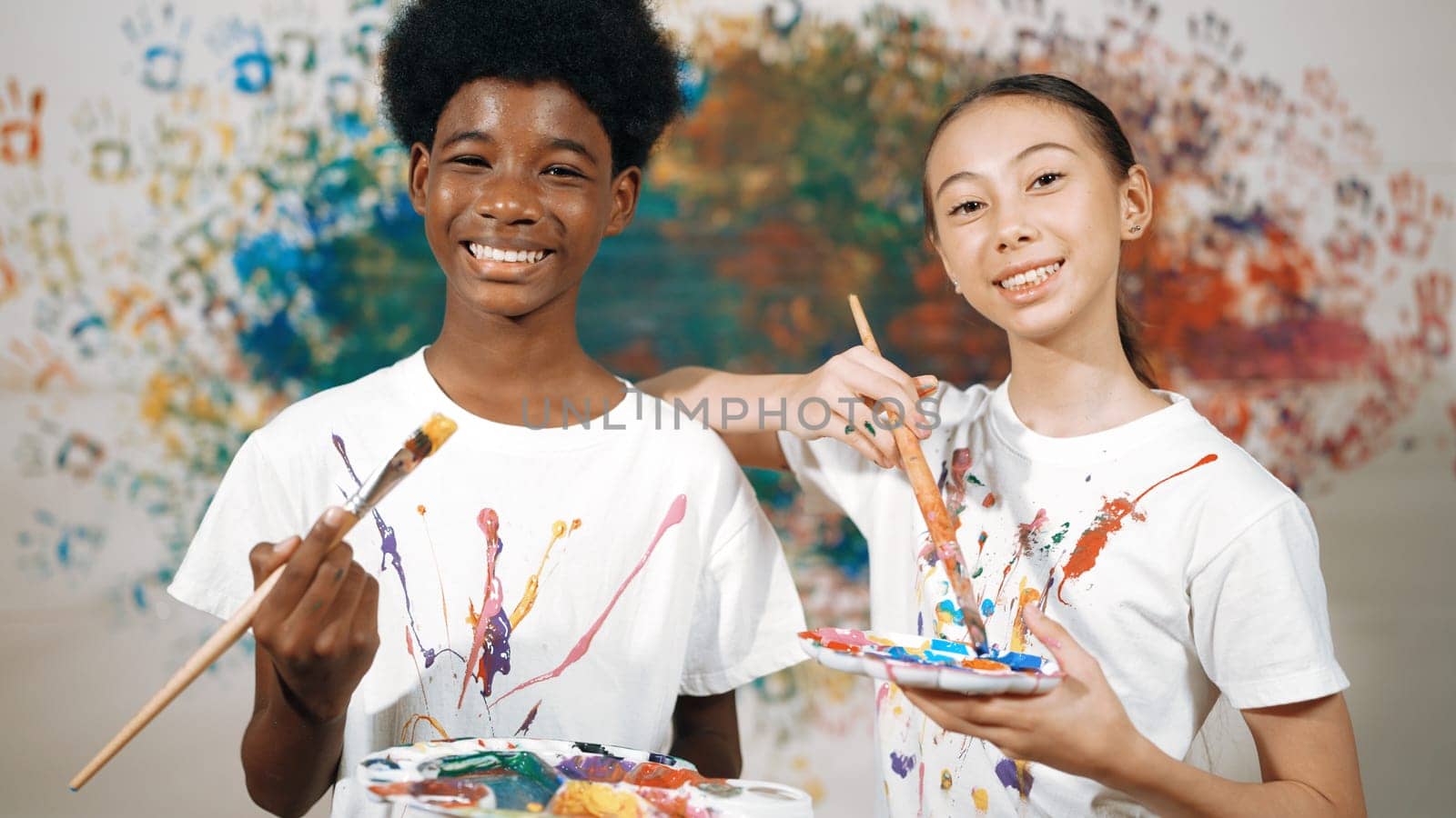 Highschool student with mixed races standing and looking at camera at art lesson. Diverse children hold the paintbrush while pose with confident at colorful stained wall. Creative style. Edification.