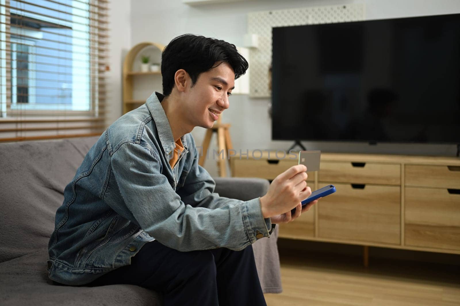 Smiling young Asian man with credit card making financial transaction on smart phone.