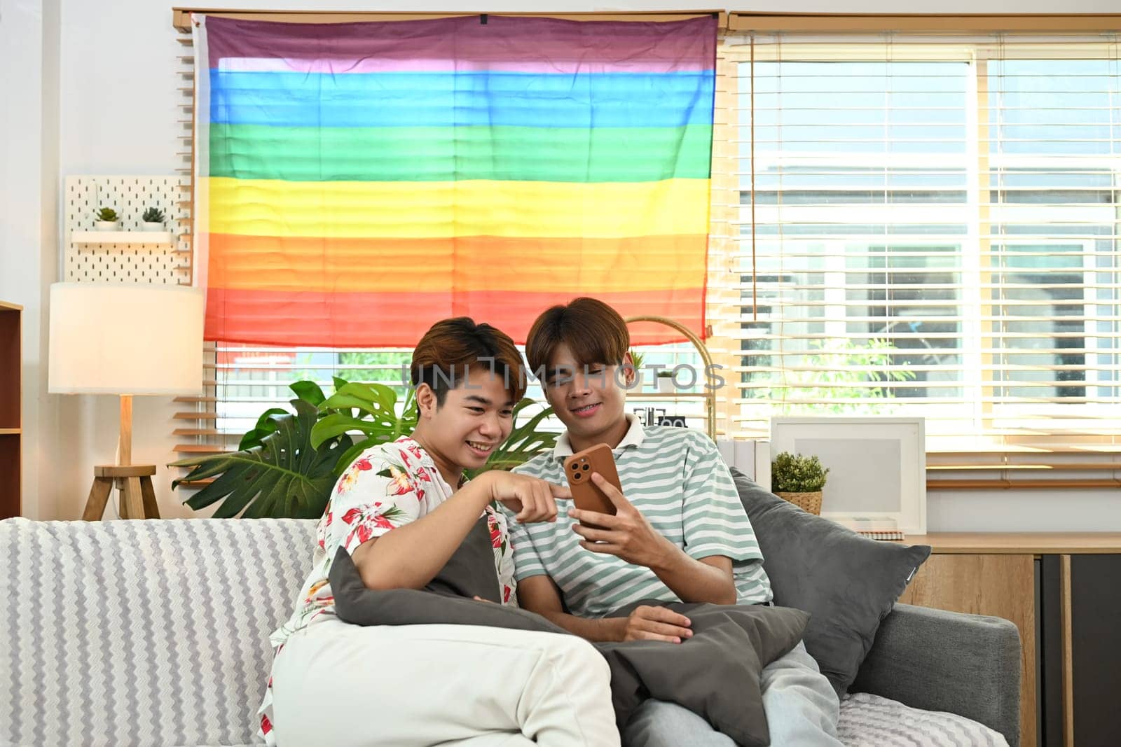 Loving young gay couple using mobile phone on sofa enjoy spending weekend together.