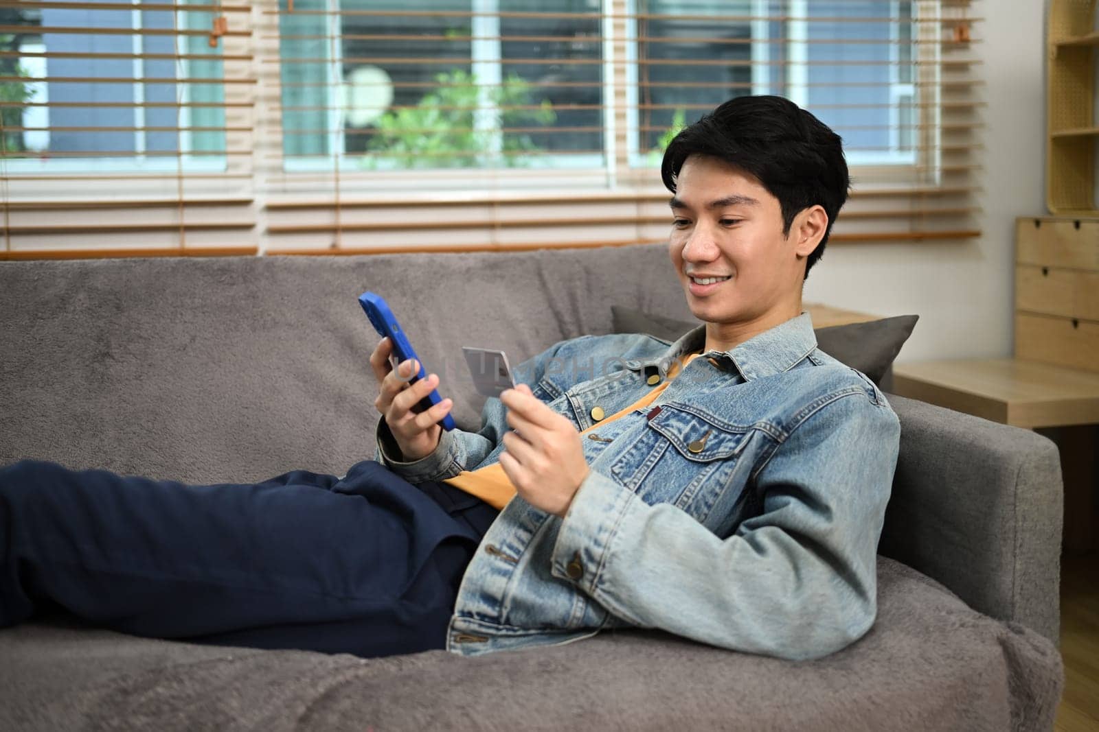 Pleased young adult man holding credit card and using mobile application for online shopping.