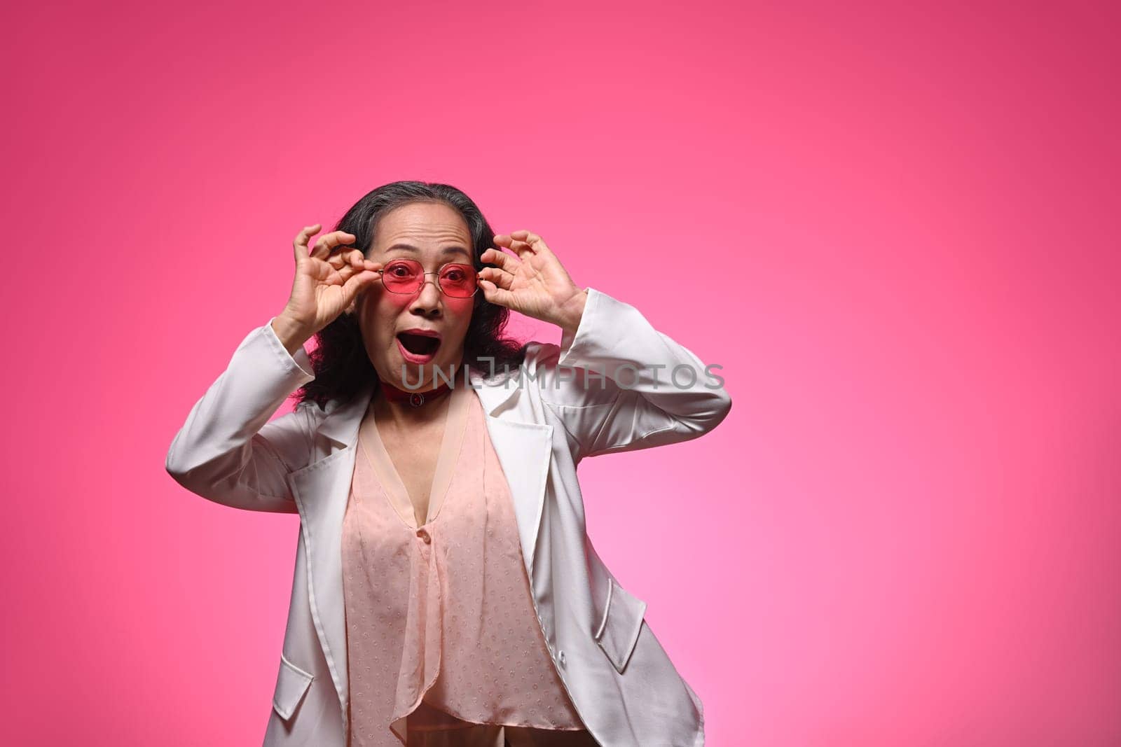Surprised elderly female in sunglasses and stylish clothes posing over pink background with copy space.