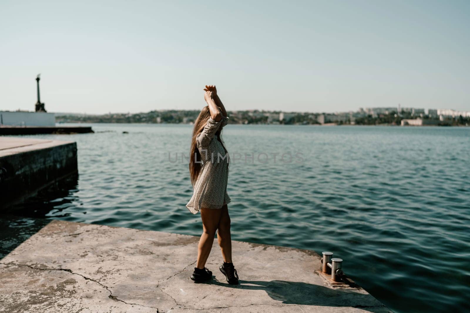 A woman is standing on a dock by the water, wearing a dress and a pair of black shoes. She is smiling and she is happy