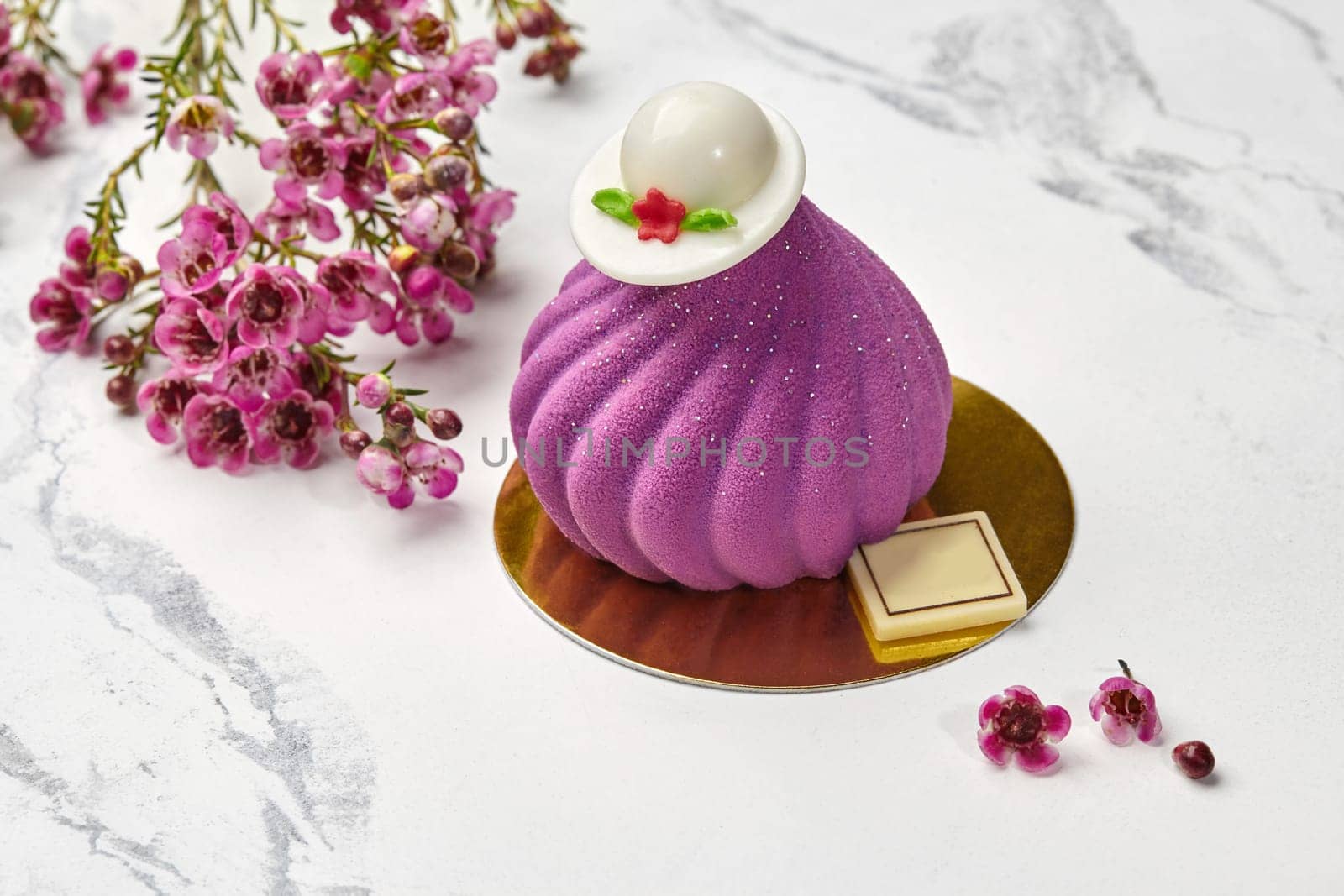 Exquisite violet swirl-shaped desserts with white chocolate emblem and decorative caps on top on light marble surface with fresh delicate spring flowers