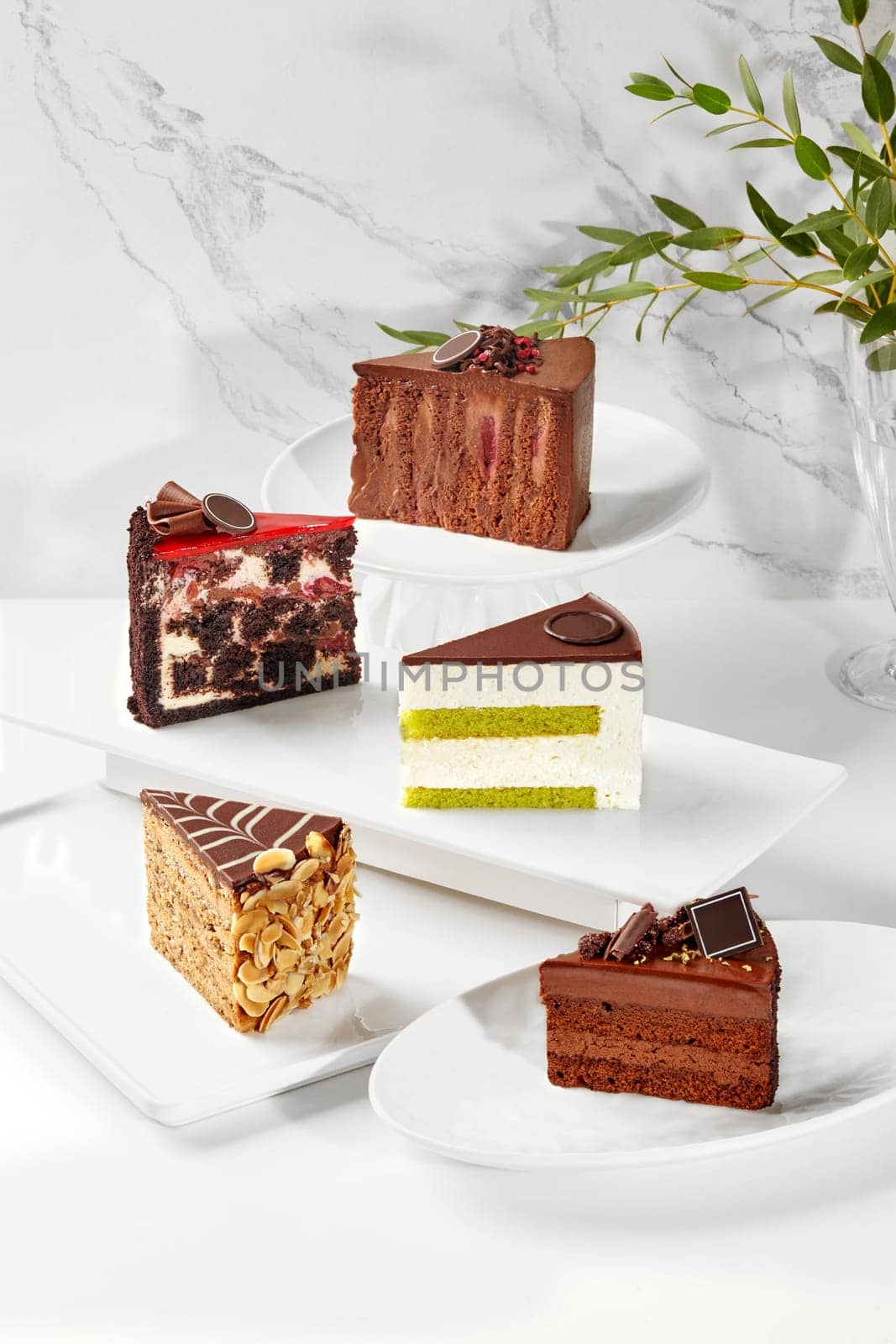 Variety of exquisite artfully decorated cake slices with chocolate and pistachio sponge, light creamy mousse, nuts and berries, presented on white plates with marble backdrop. Popular baked sweets