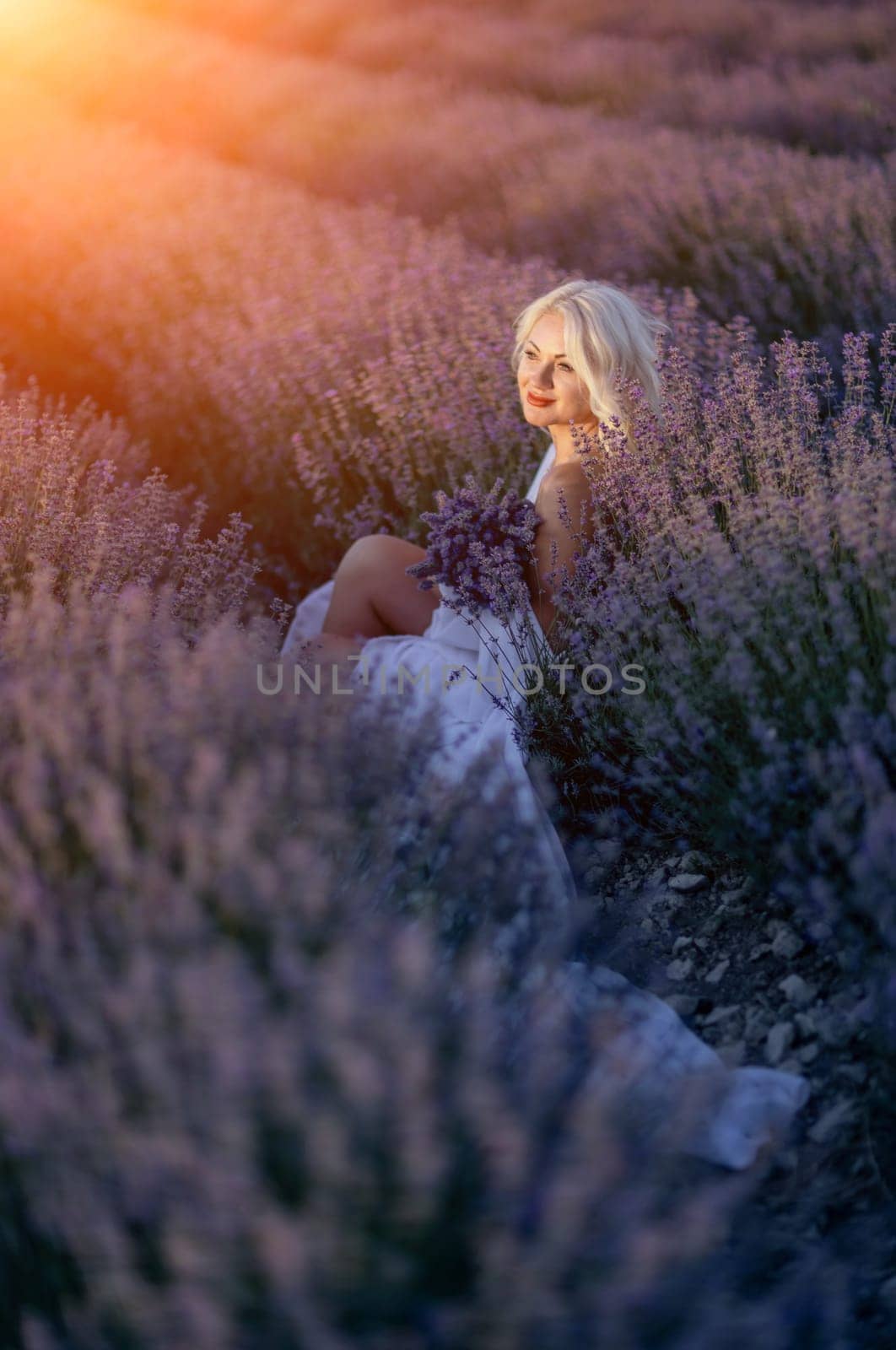 Blonde woman poses in lavender field at sunset. Happy woman in white dress holds lavender bouquet. Aromatherapy concept, lavender oil, photo session in lavender.