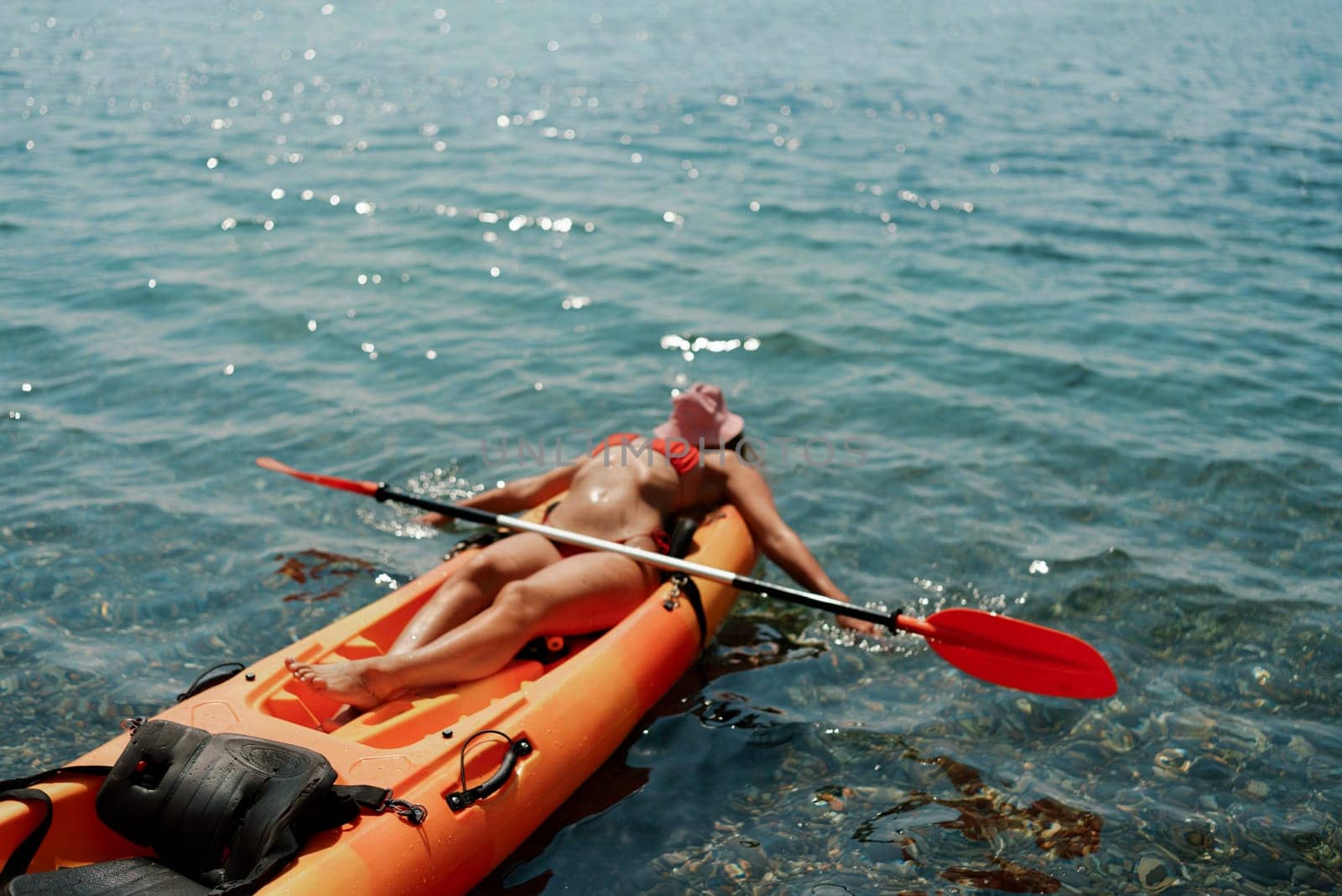 A woman is laying on a kayak in the water. The kayak is orange and has a paddle on it. The woman is wearing a red hat