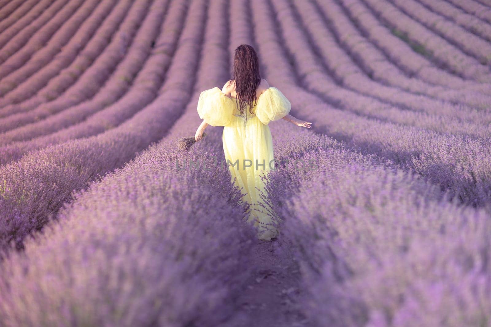 A woman in a yellow dress walks through a field of lavender. The field is full of purple flowers, and the woman is enjoying the scenery