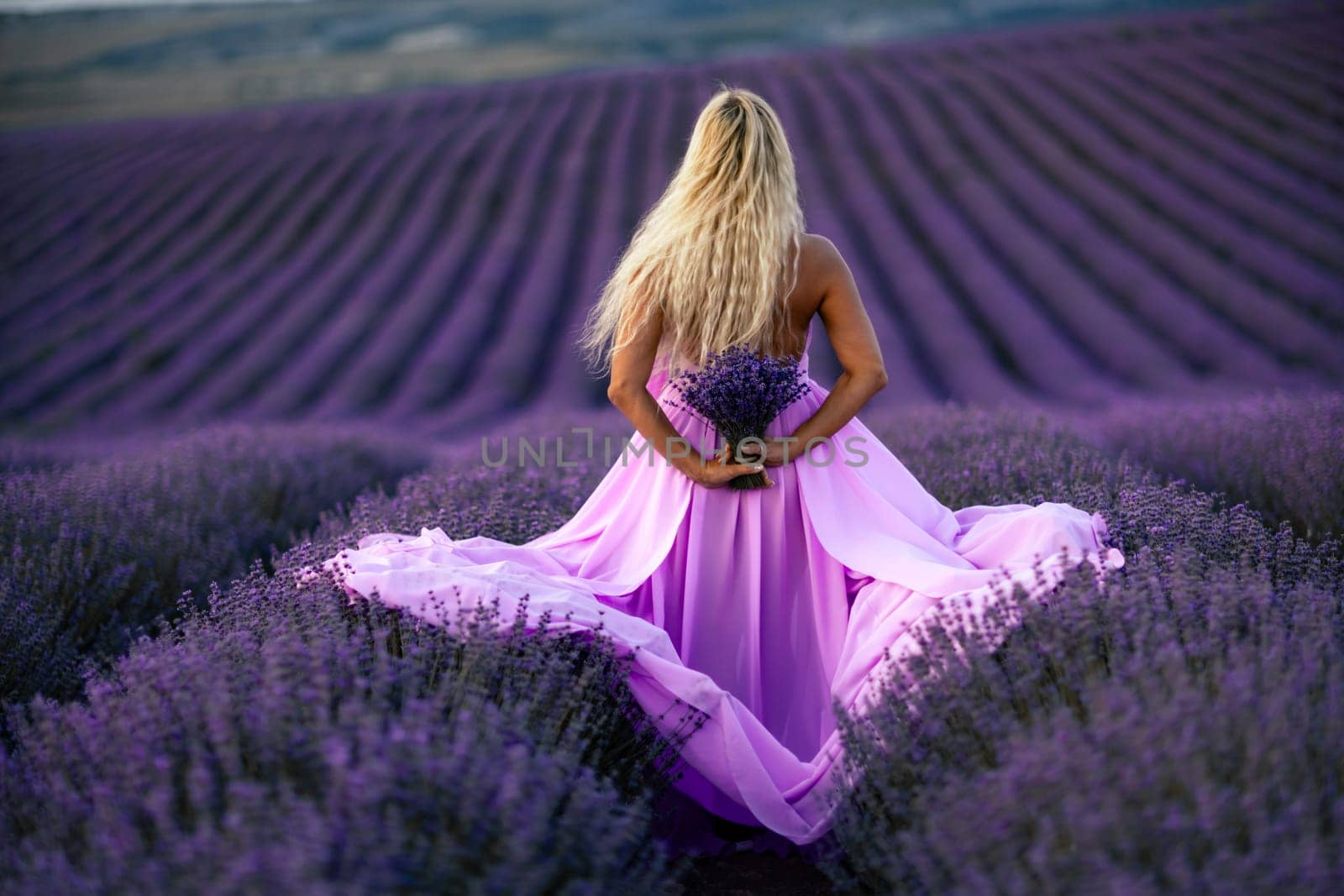 Back view woman lavender sunset. Happy woman in pink dress holds lavender bouquet. Aromatherapy concept, lavender oil, photo session in lavender.