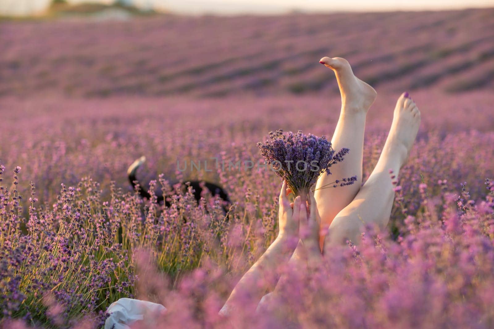 A woman is laying in a field of purple flowers with her legs spread out. She is holding a bouquet of flowers in her hands. The scene is peaceful and serene