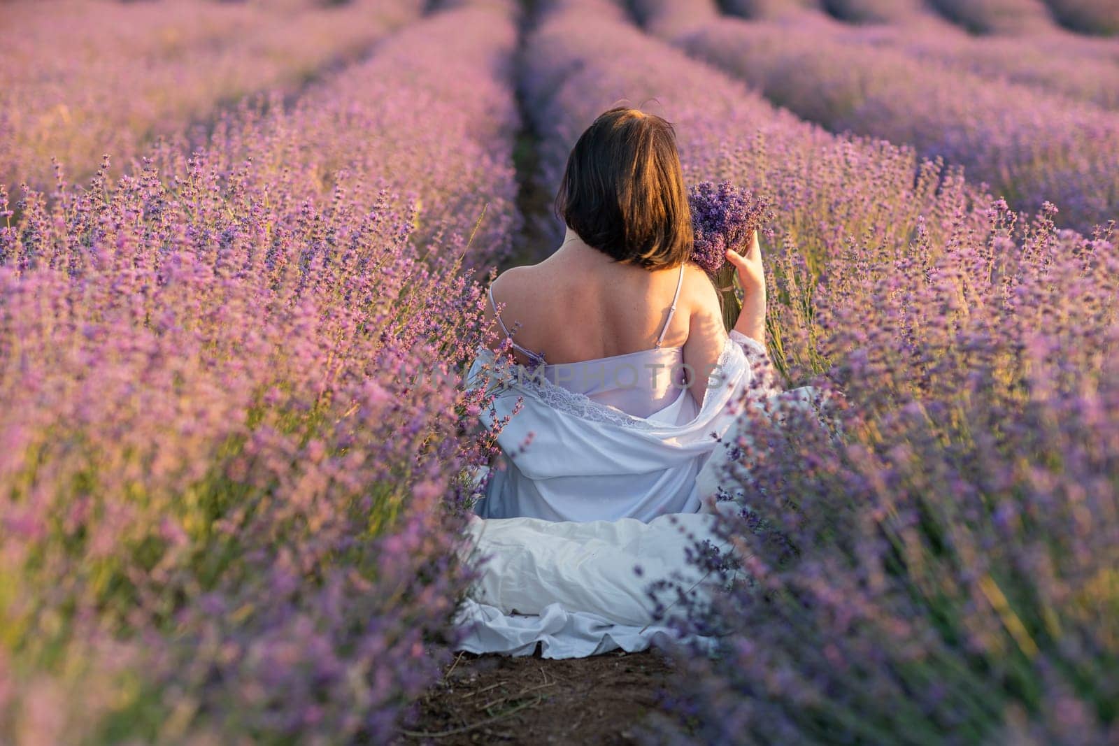 A woman sits in a field of lavender flowers. She is holding a bouquet of flowers in her hands. The scene is serene and peaceful, with the woman surrounded by the beauty of nature