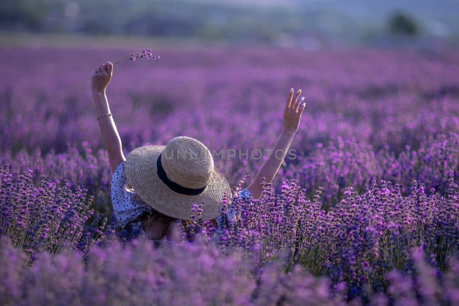 A woman is in a field of purple flowers, wearing a straw hat and holding a flower. Scene is peaceful and serene, as the woman is surrounded by the beauty of nature