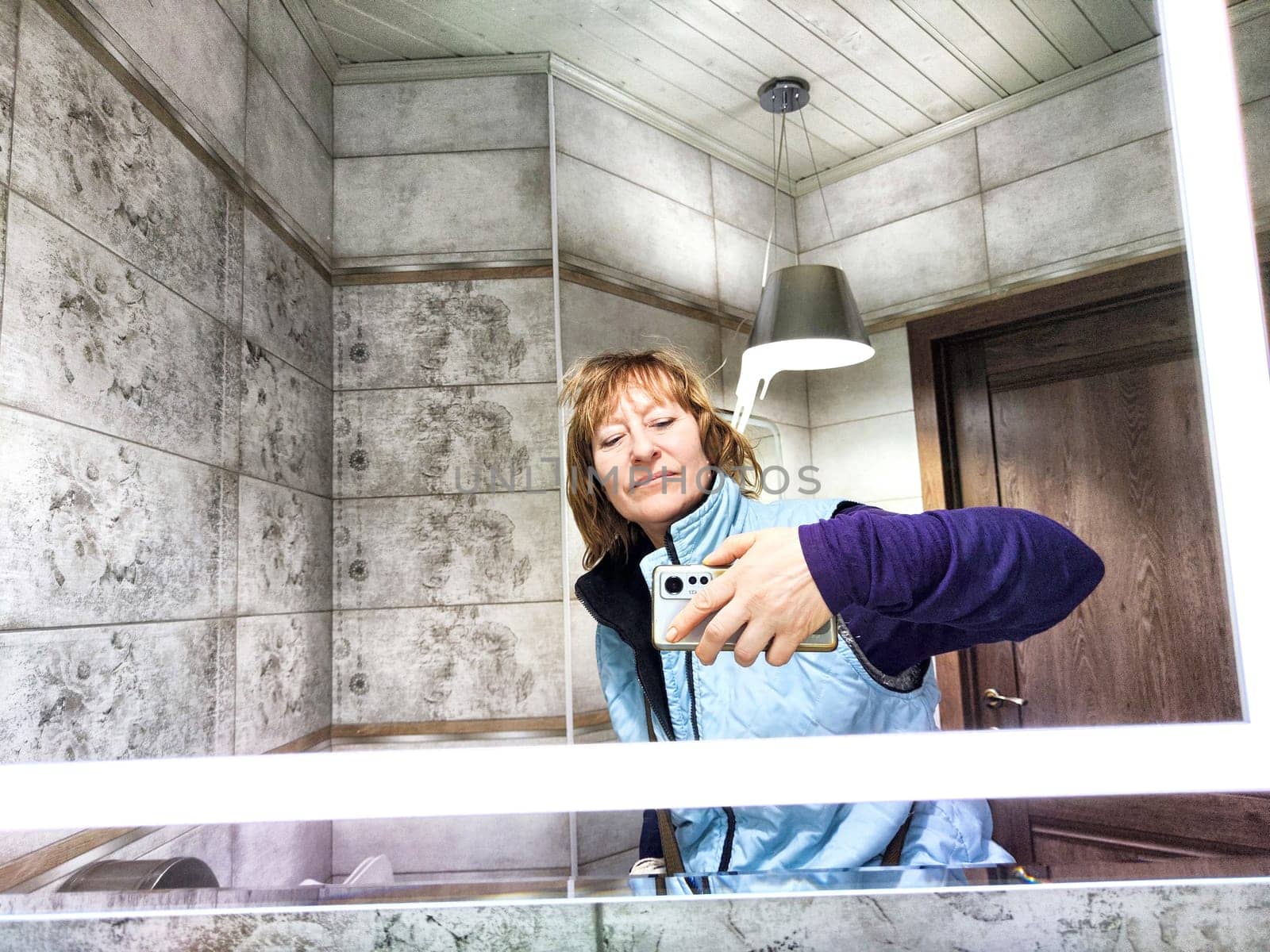 Mature woman captures her reflection while taking selfie. The reflection of middle aged girl with a phone in the mirror