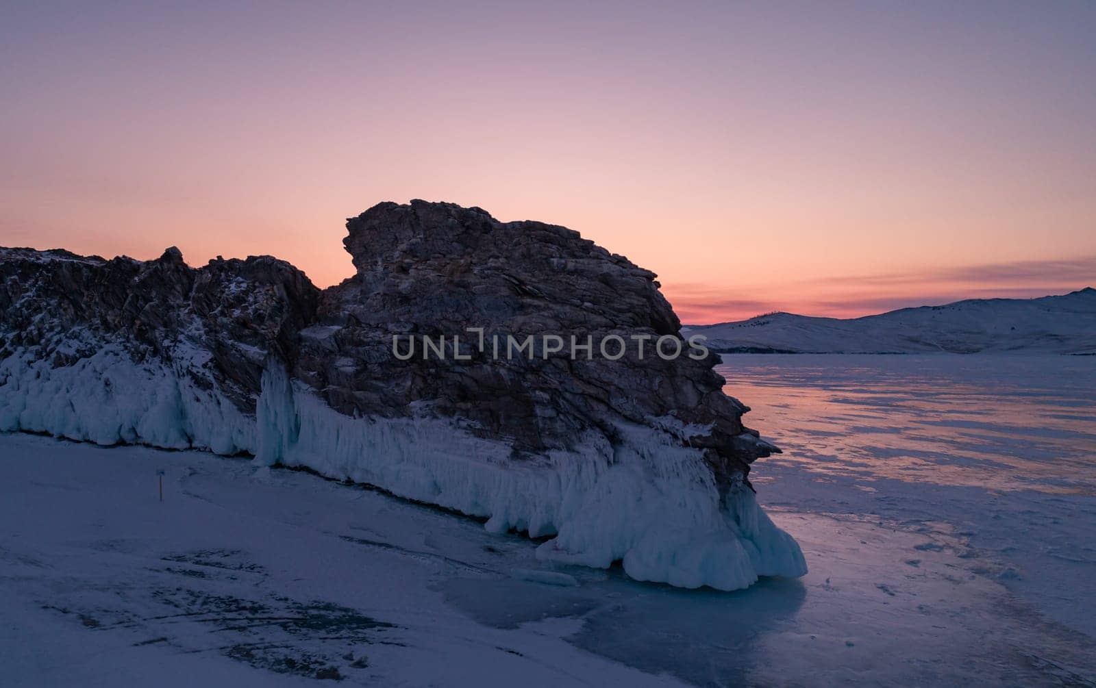 Aerial over the rocky island in lake Baikal. Winter landscape of frozen Baikal at beautiful orange sunrise. Sun reflections on the ice. Popular tourist spot.