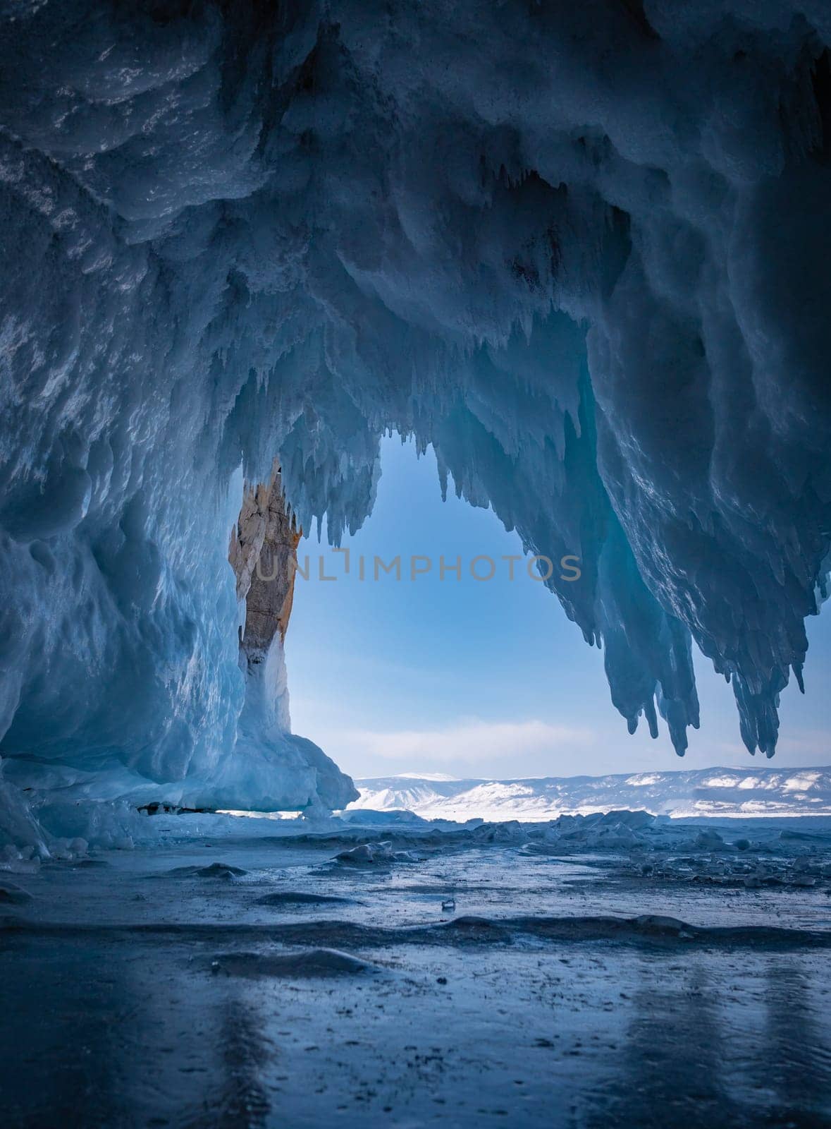 Inside a stunning ice cave on Lake Baikal, large icicles hang from the ceiling, creating a breathtaking winter landscape. Snow-covered mountains can be seen far in the distance.