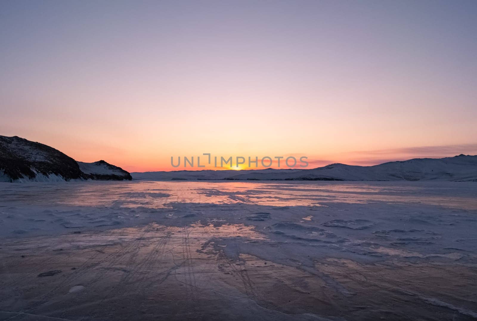 Snowy cracky ice of lake Baikal and the rocky island. Winter landscape of frozen Baikal at beautiful lilac sunrise. Sun reflections on the ice. Popular tourist spot by Busker