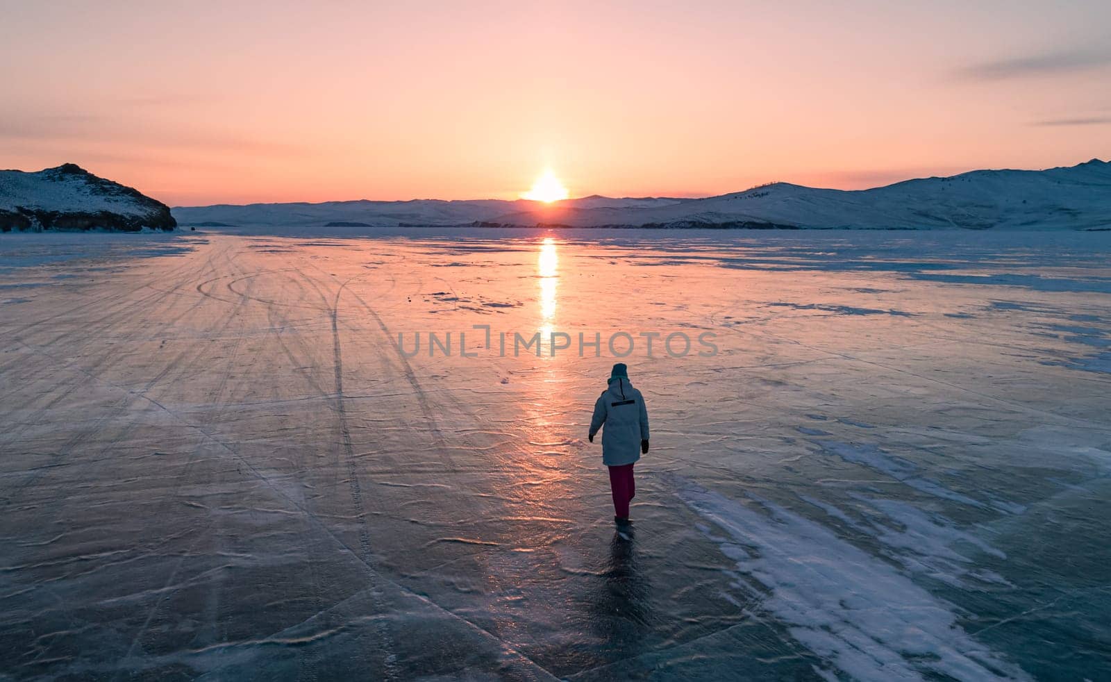 Aerial view on the young woman walking on the blue cracked ice of Baikal at beautiful orange sunrise. Sun reflections on the ice. Winter landscape of frozen Baikal by Busker