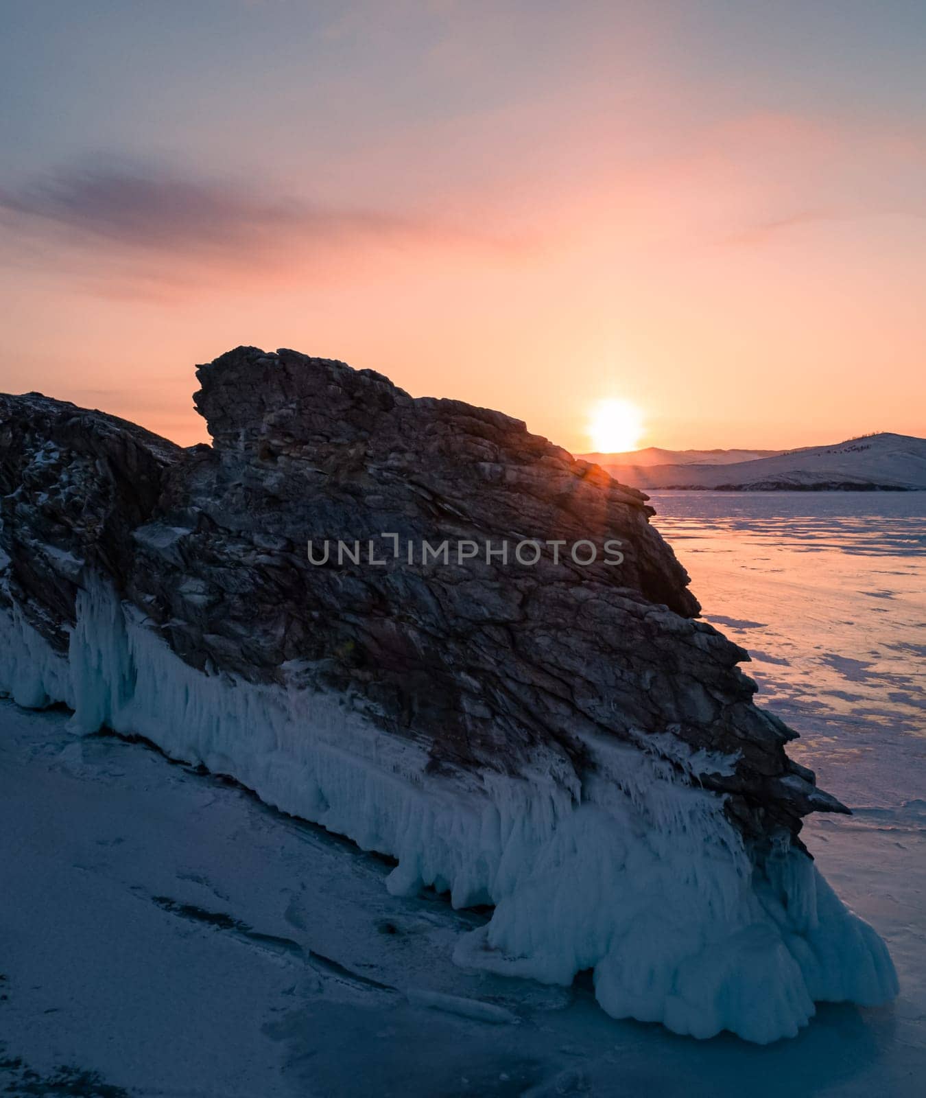 Aerial over the rocky island in lake Baikal. Winter landscape of frozen Baikal at beautiful orange sunrise. Sun reflections on the ice. Popular tourist spot by Busker