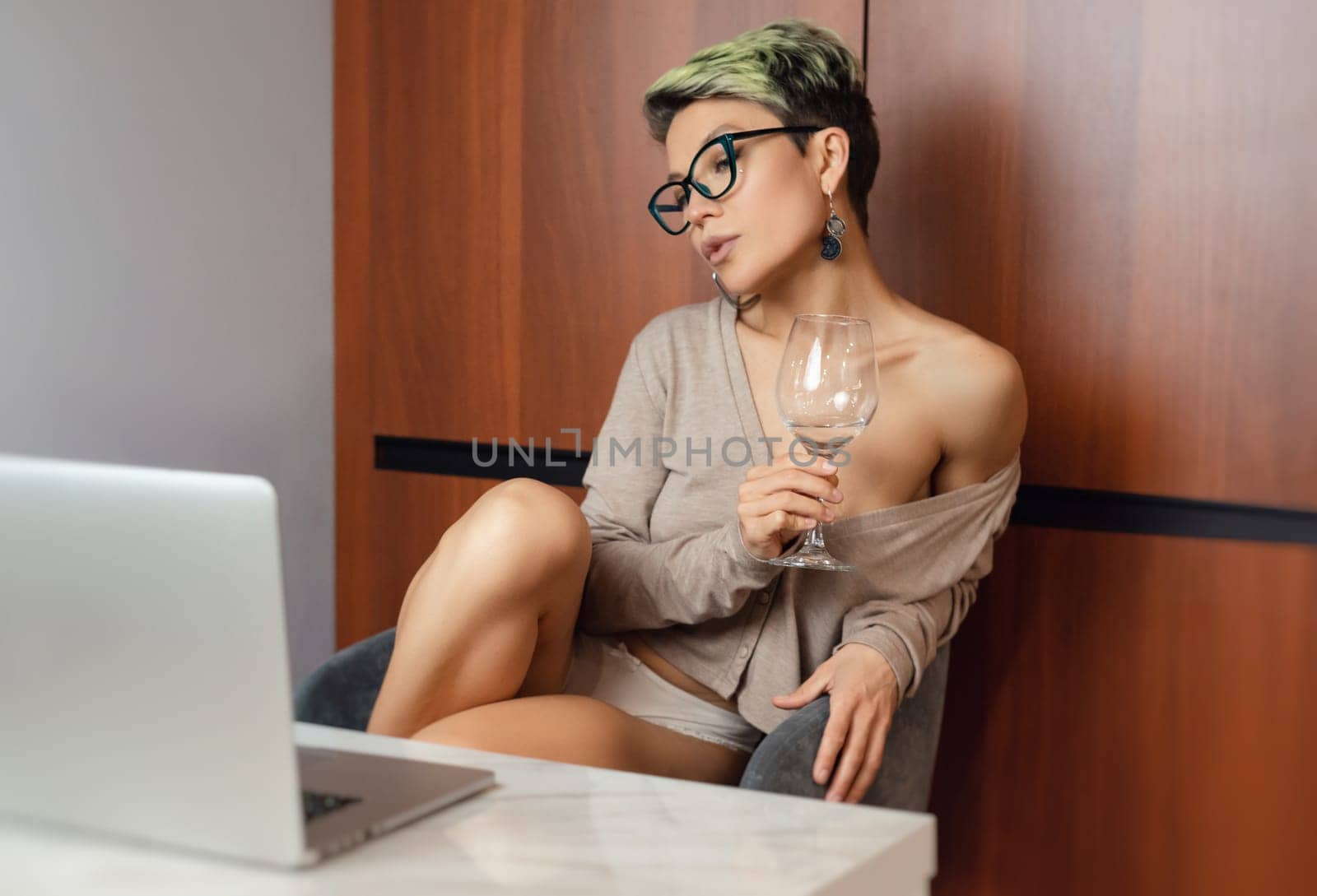 a beautiful girl with short hair and glasses is sitting indoors at a laptop, with her feet on the table, chatting and working online with a glass of water