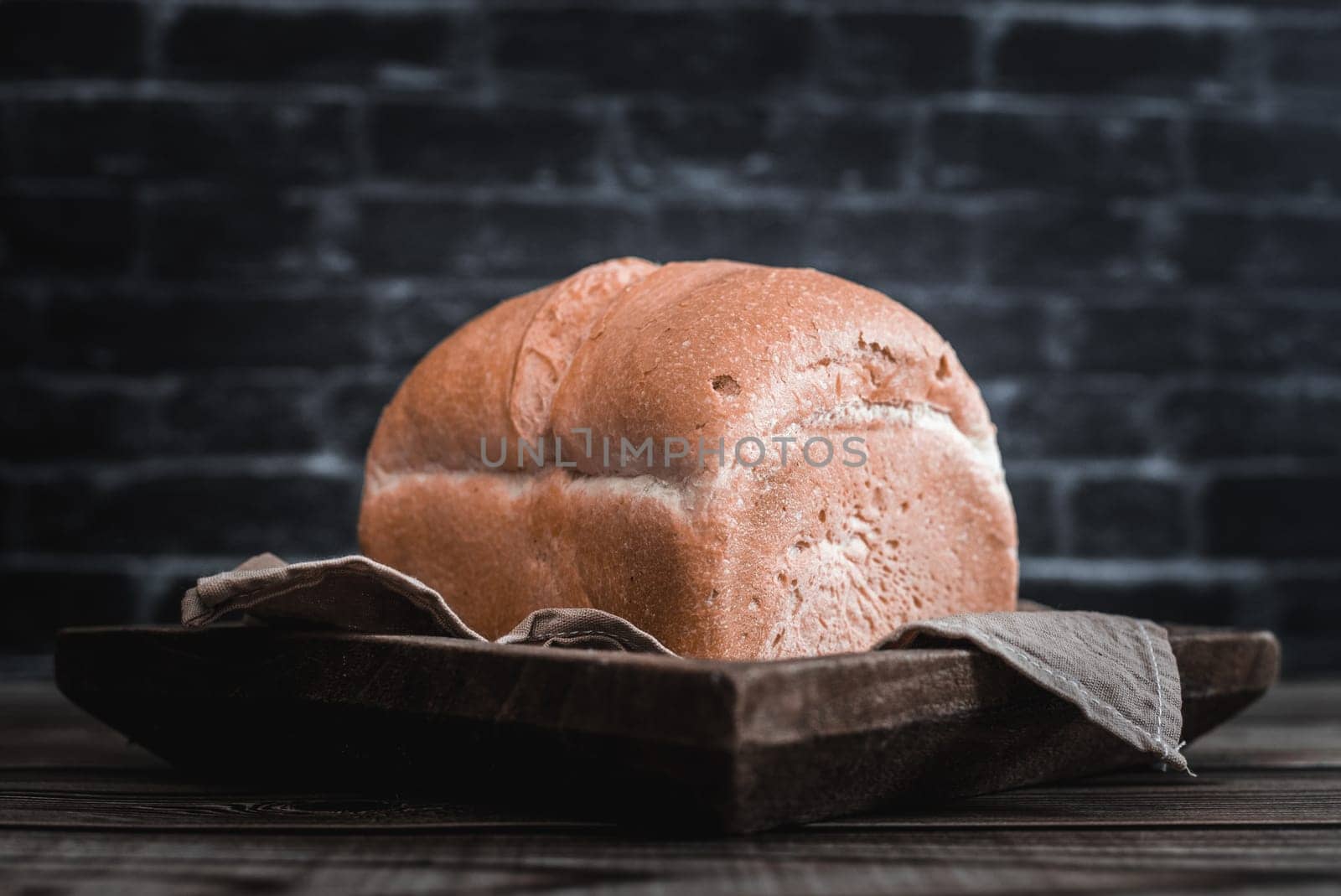 One square white bread in a wooden dish with a kitchen napkin lies on a brown wooden table against a blurred black brick wall, close-up view from below. The concept of baking bread.
