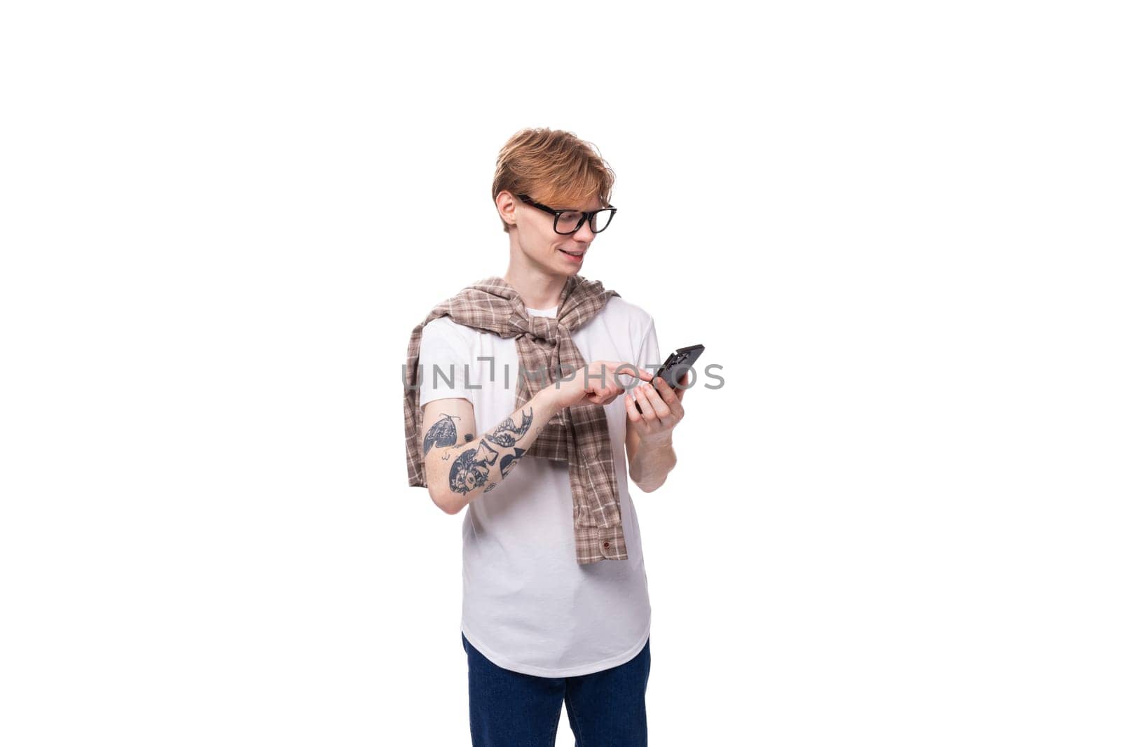 cute red-haired guy in glasses in a summer shirt writes a message in the phone on a white background with copy space.