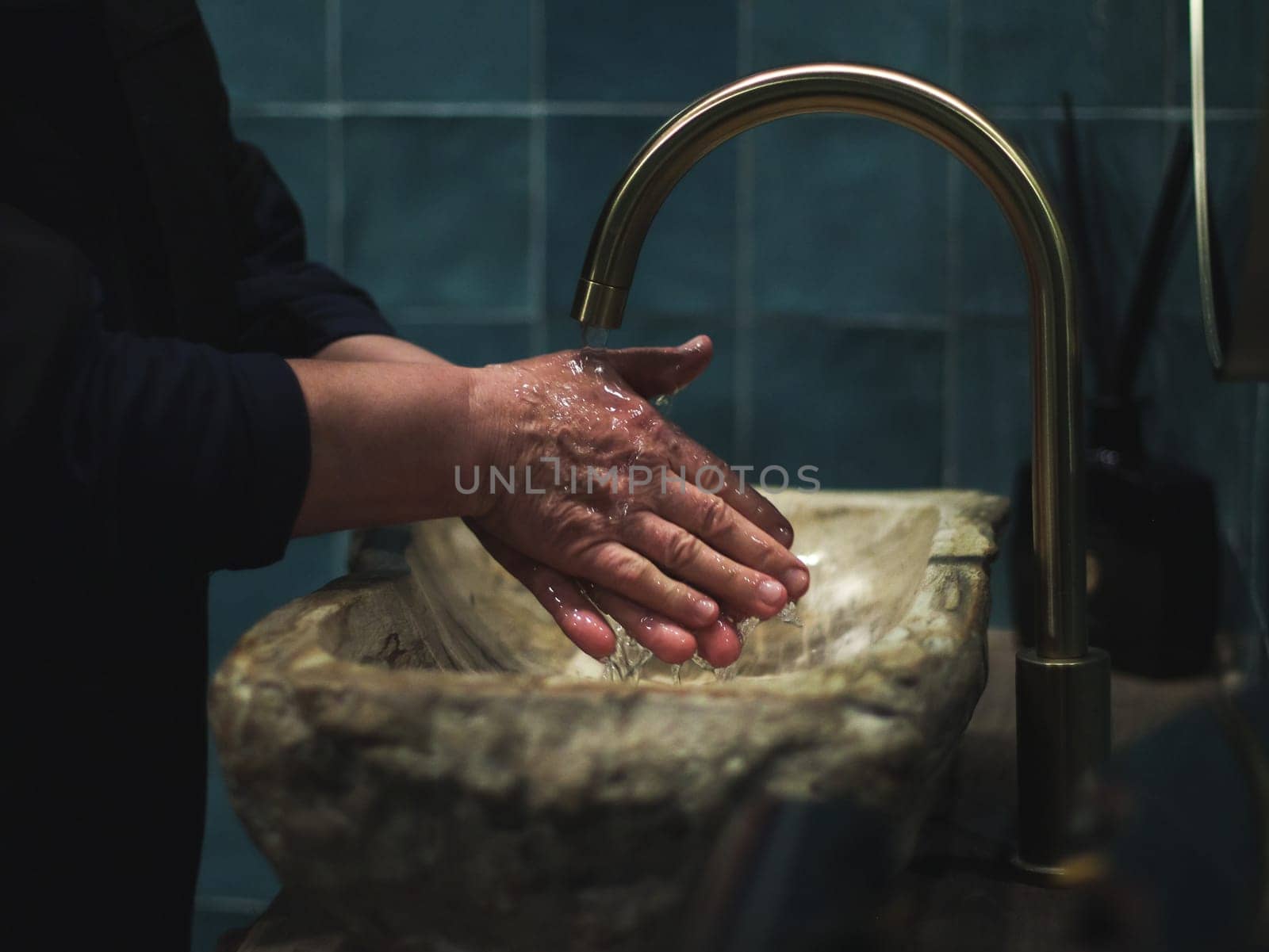 Elderly senior lady in black clothes washes her hands under the tap in the stone washbasin of the public toilet in the restaurant, close-up side view. Seniors body part concept, personal hygiene, dark style, bathroom interior.